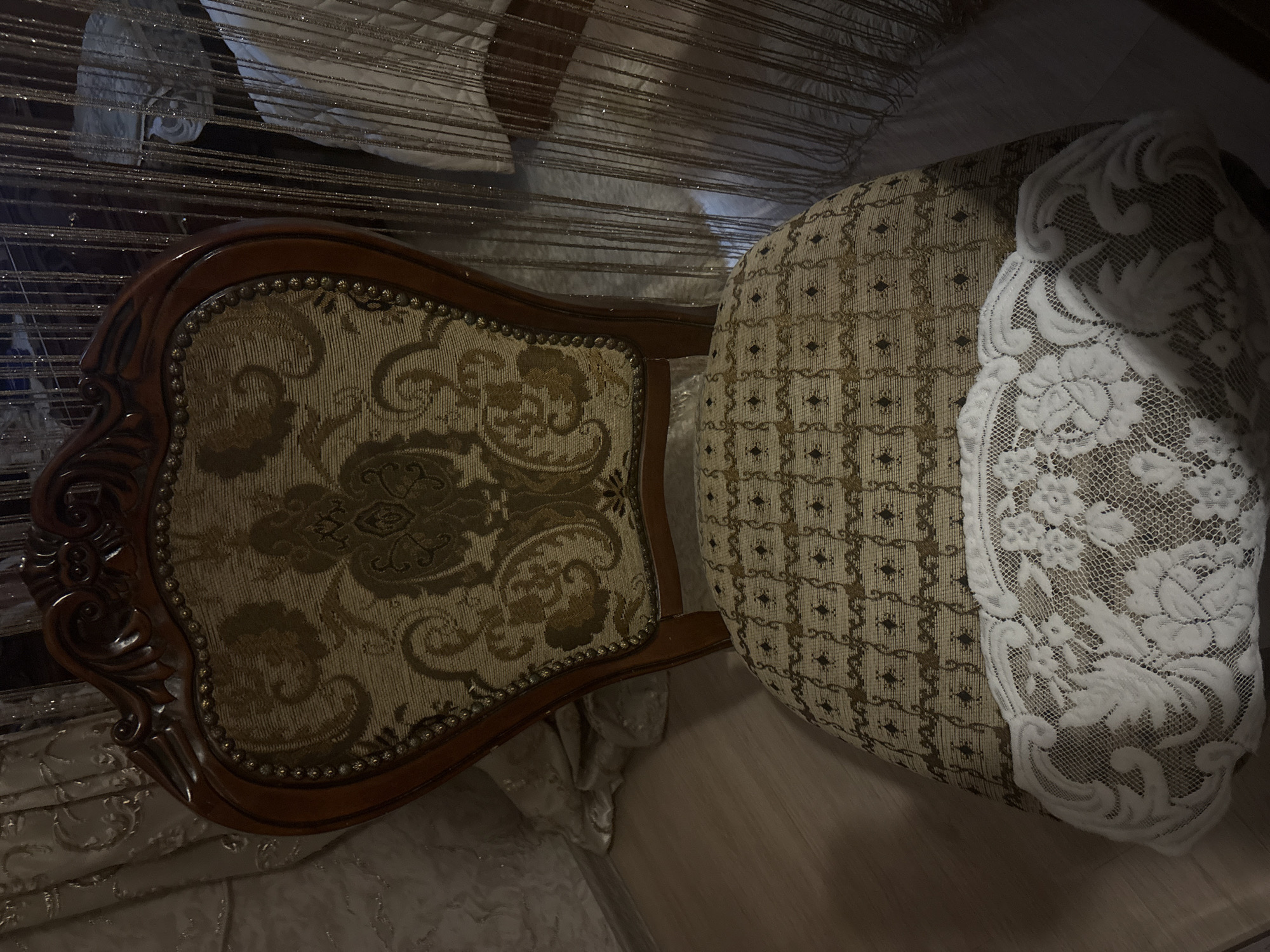 The image shows a close-up, high-angle view of an ornate chair. The chair has a wooden frame with a decorative pattern and is upholstered in a floral tapestry fabric. The seat of the chair is covered with a patterned fabric and partially covered by a white lace cloth. The background includes a curtain or wall hanging with shimmering strands. There is insufficient information to determine the location in Krasnoyarsk, Russia.