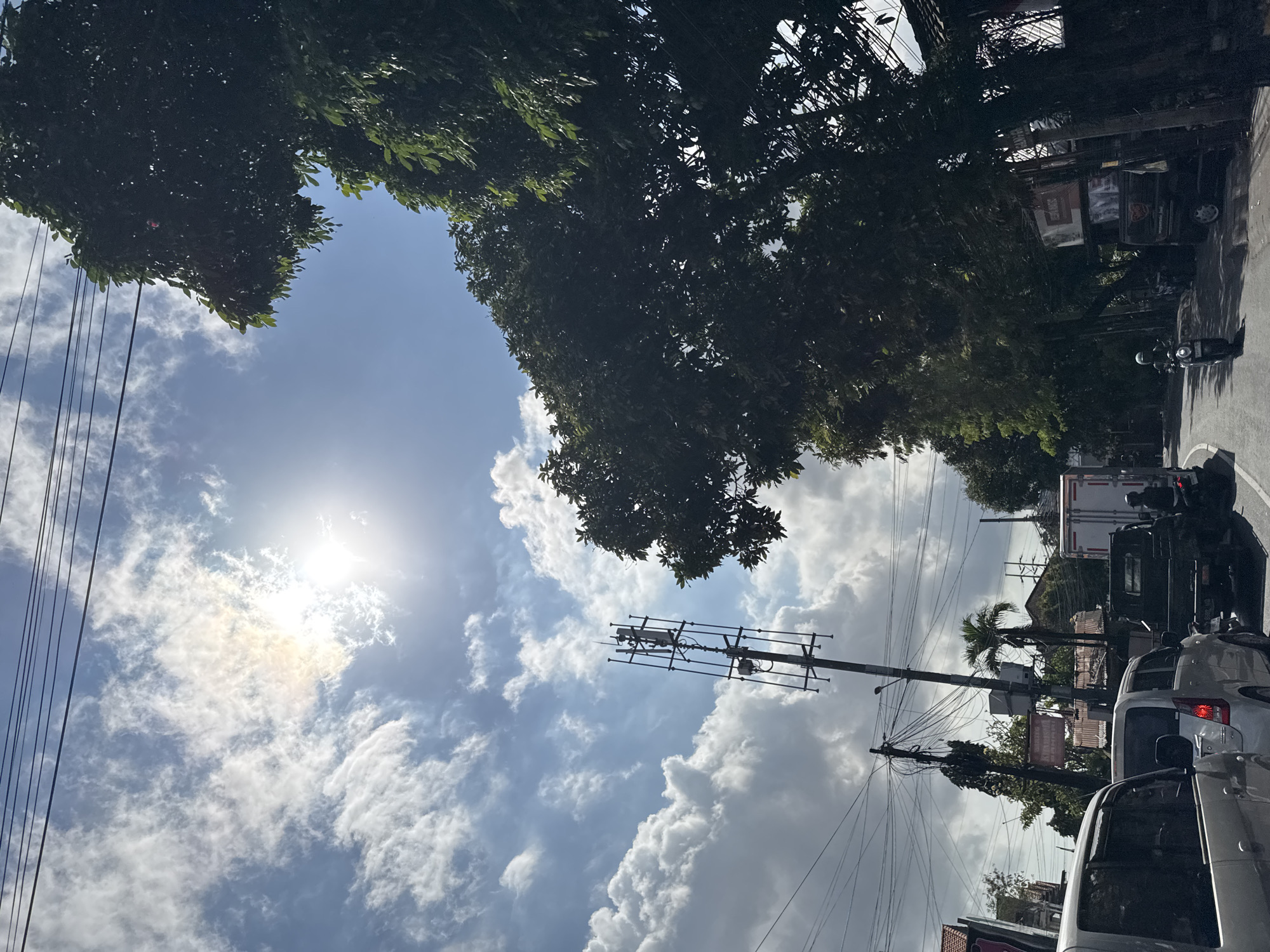 The image depicts a street scene in Denpasar, Indonesia, likely during daytime given the bright sun and clear sky. The perspective is from within a vehicle, suggesting it is a road in a city.
In the foreground, several vehicles are visible, including the rear of a silver car on the left and a white van directly behind it. Further down the road, a dark-colored truck is stopped, with a person on a motorcycle behind it. To the right, a motorcycle with a rider wearing a helmet is moving along the road.
Dominating the middle ground and background are several tall structures and natural elements. A cell tower, a prominent metal lattice structure, stands tall on the left side of the road. Next to it, telephone poles with a dense network of wires are visible, indicating urban infrastructure. Lush green trees with dense foliage line the right side of the street, creating a canopy over parts of the road and sidewalk. Palm trees can be seen in the distance behind the cell tower. Buildings with reddish roofs are visible behind the trees and infrastructure.
The sky is bright blue with scattered white clouds, and the sun is shining intensely, creating some lens flare. The overall impression is of a busy urban street in a tropical climate.