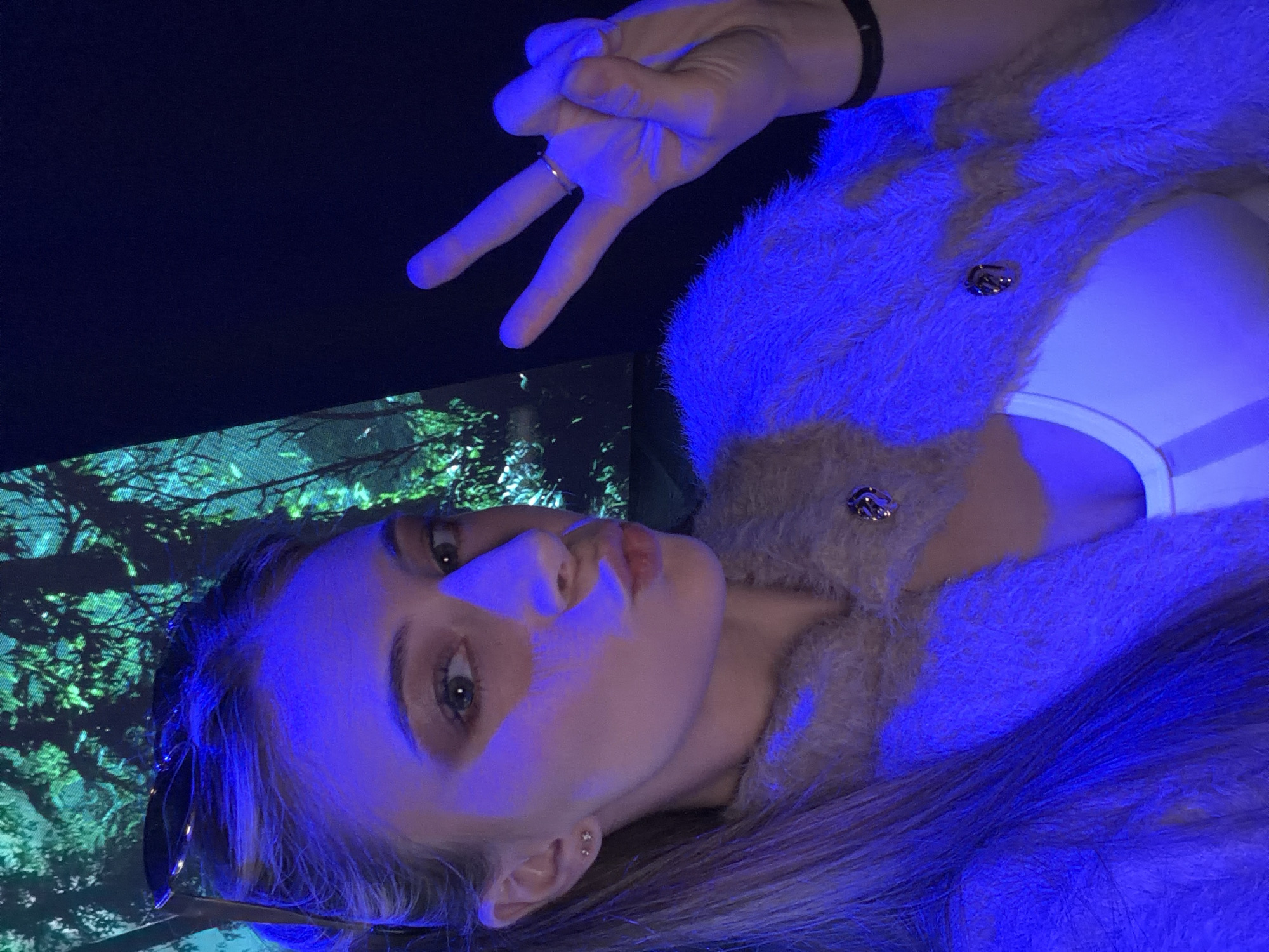 The photograph is a selfie of a young woman with blonde hair, shot indoors with a prominent blue lighting effect. She is wearing a fuzzy beige cardigan with ornate silver buttons, and a white top underneath. She is making a peace sign with her right hand. In the background, there is a screen or projected image depicting a forest scene. The presence of the screen displaying a forest scene might indicate the location is an art installation or an interactive museum experience. Without further information or visual clues specific to Plovdiv, Bulgaria, it is impossible to definitively pinpoint the exact location.
