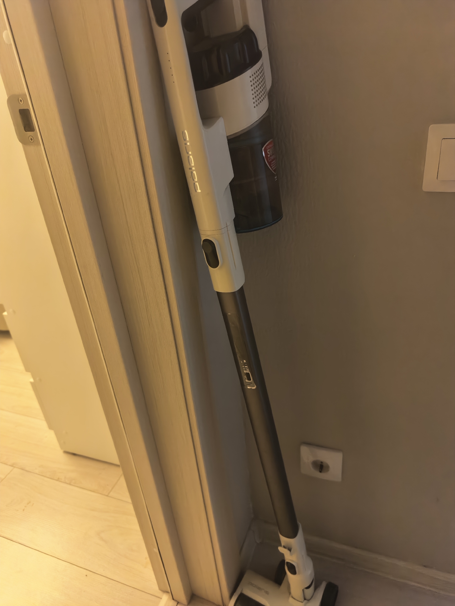 A white stick vacuum cleaner is leaning against a light gray wall, positioned to the right of a white door frame. The vacuum has a dark gray tube and a white body with a transparent dustbin. The brand name "POLARIS" is visible in silver lettering on the vacuum's tube. To the right of the vacuum, a white electrical outlet is mounted on the wall. The floor is a light-colored wood laminate. The scene is indoors, with artificial lighting creating some shadows.