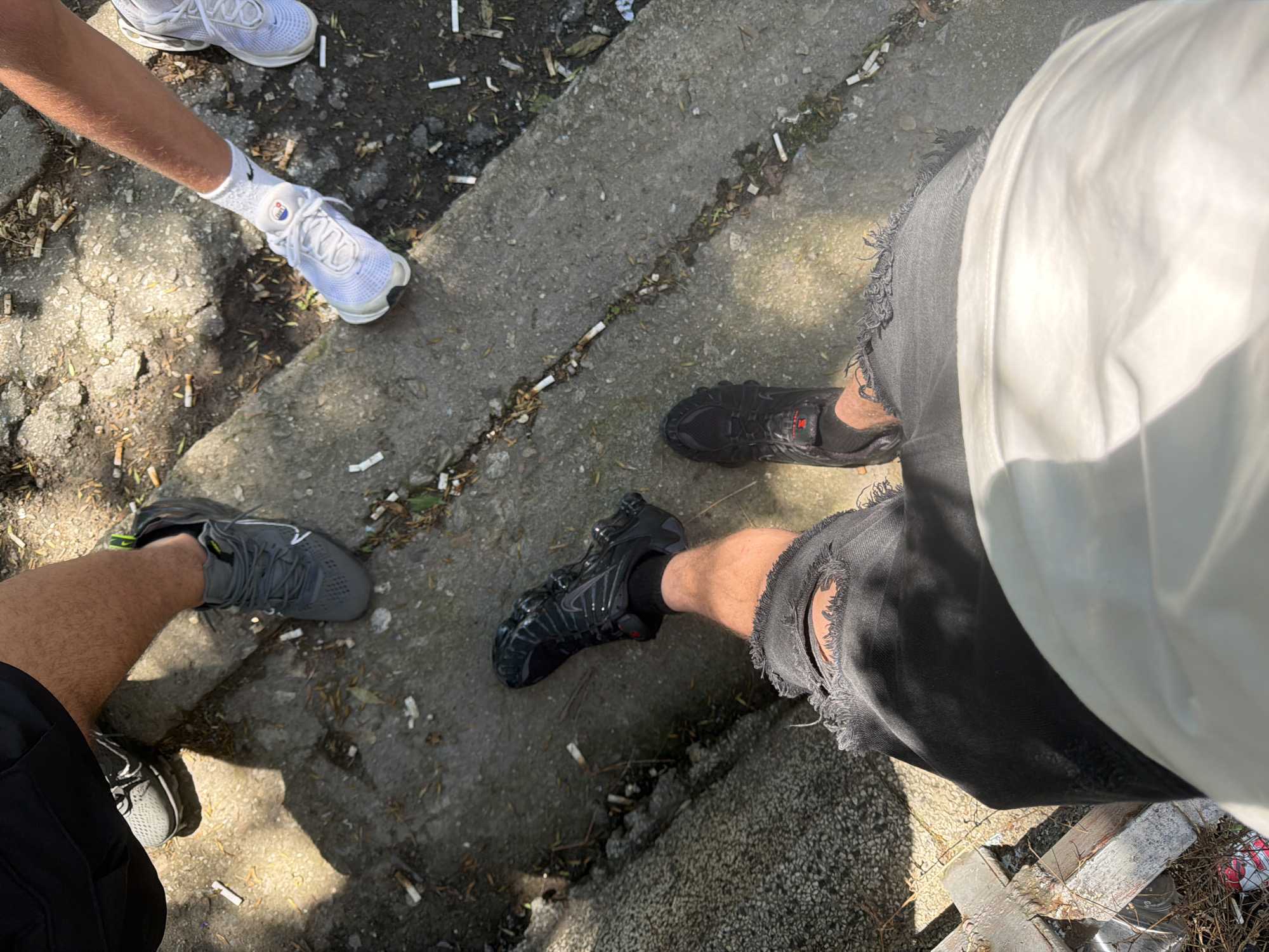 The image captures an overhead perspective of several individuals' legs and feet, situated on a paved surface littered with cigarette butts. In the lower right, one person wears black ripped shorts and black athletic shoes. To the left and slightly above, another individual is seen with their legs bare from the thigh down, clad in black ripped shorts and black athletic shoes with red accents. Further up and to the left, a third person's legs are visible, wearing dark shorts and grey athletic shoes. On the right side of the frame, a fourth person's legs are shown, wearing white socks and white athletic shoes. The ground appears to be concrete or asphalt, with patches of dirt and some sparse greenery. The lighting suggests it is daytime. No specific landmarks in Plovdiv, Bulgaria, are identifiable from this image.