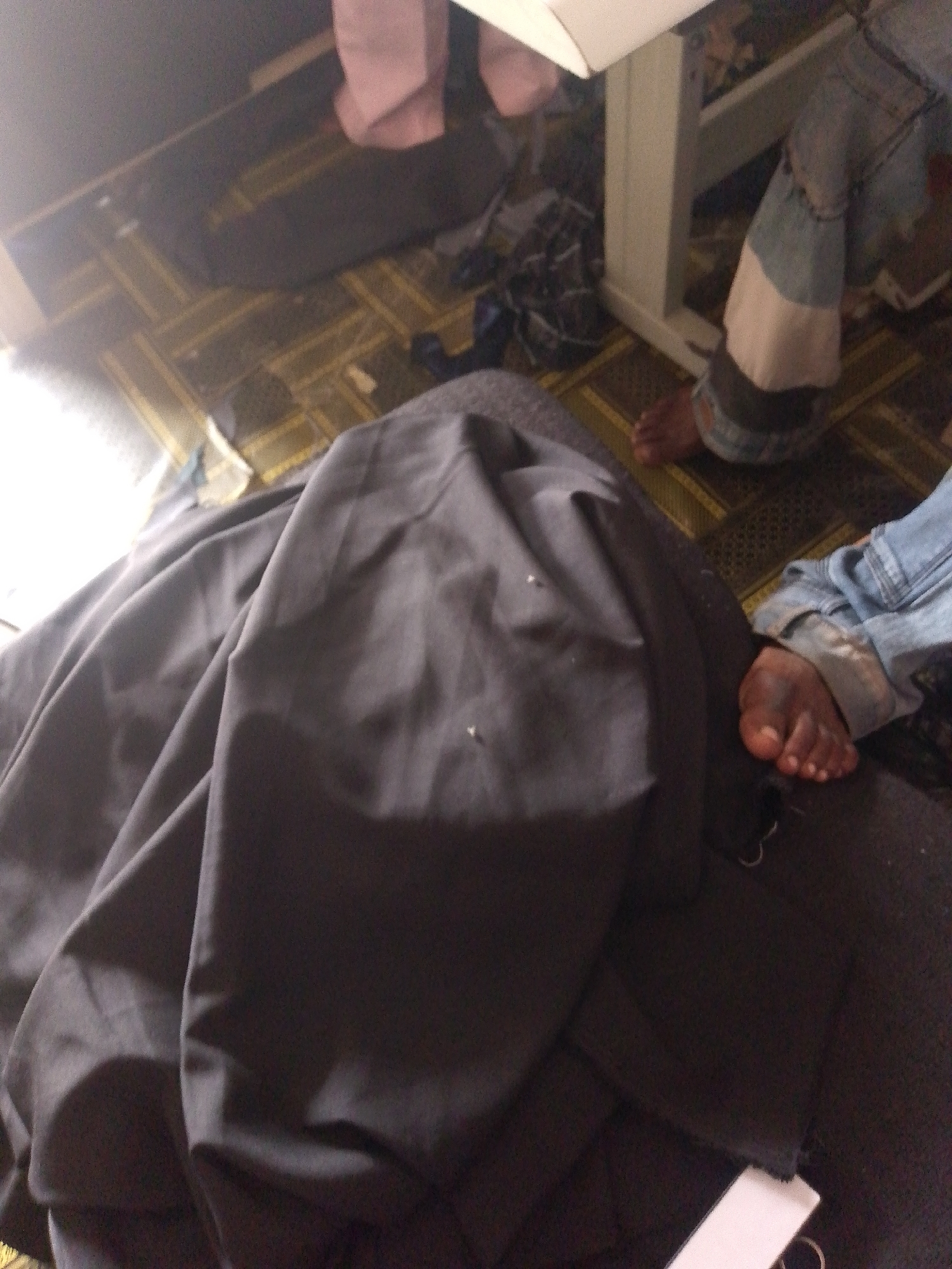 In Jauro Yinu, Nigeria, a low-angle, medium shot captures a dark, wrinkled fabric draped over what appears to be a human form. The fabric dominates the foreground, obscuring most details. To the right of the fabric, a person's legs are visible, clad in light blue denim jeans with patches of white and grey fabric. One foot, with dark skin, is bare and rests on a patterned floor. The background suggests an indoor setting with a wooden table or desk and a blurred object with pink fabric. The lighting is dim, with a bright glare emanating from the left side of the frame. The scene appears to be an interior shot, possibly in a residential or workshop environment.