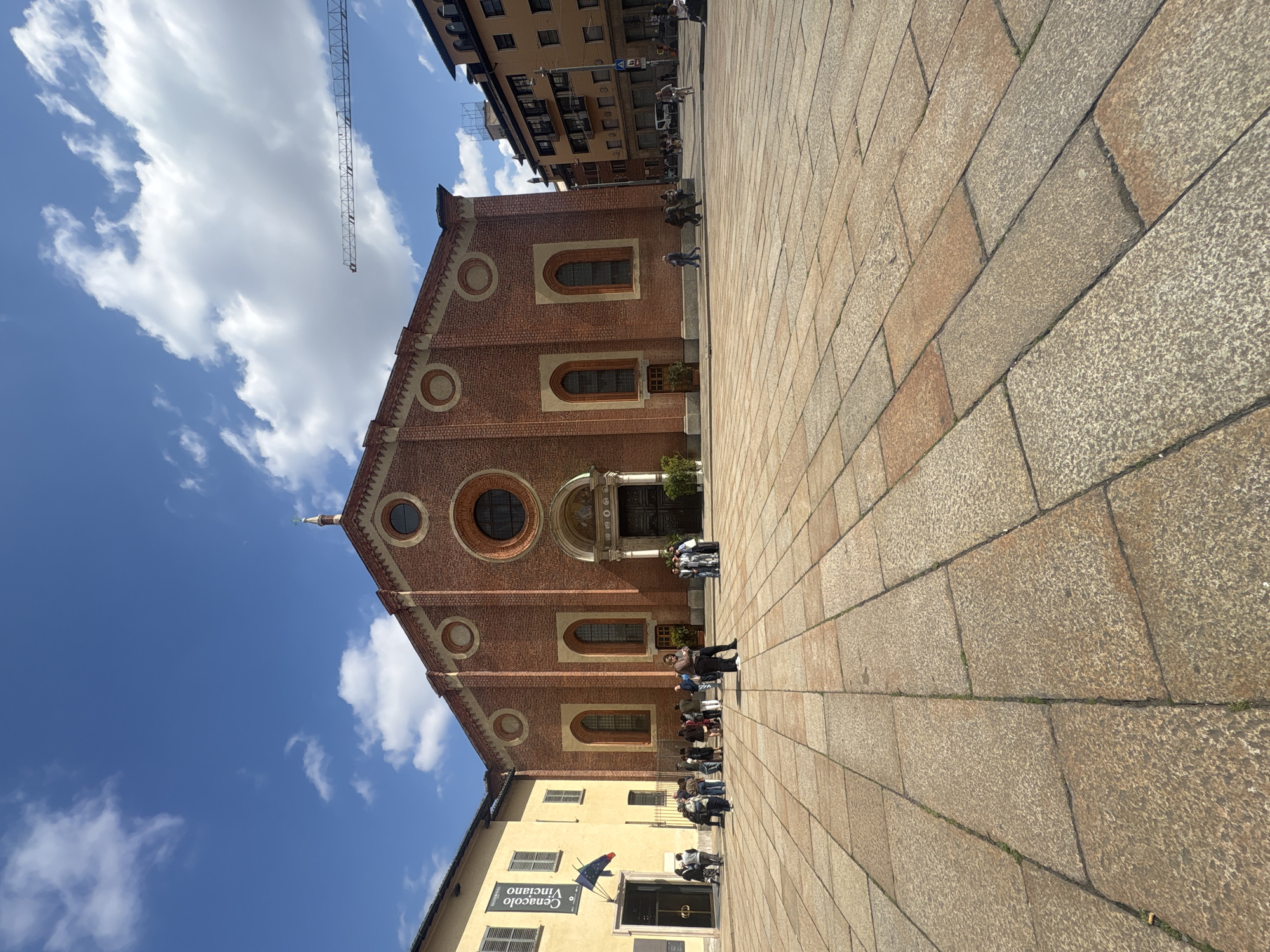 This image depicts a vibrant outdoor scene in a public square in Milan, Italy, under a clear, sunny sky with scattered white clouds. The main subject is a large, red-brick building, likely a historical church or monument, characterized by its classical architecture with multiple arched windows and prominent circular windows (oculi) on its facade. A central, ornate entrance with steps leads into the building.
Adjacent to the brick building, on the left, is a smaller, yellow-painted building with dark window shutters. A banner hangs from this yellow building, bearing the Italian text "Cenacolo Vinciano," which points to the location of Leonardo da Vinci's famous "Last Supper" fresco, specifically at the refectory of the Santa Maria delle Grazie church.
The foreground consists of an extensive paved area made of large, light-colored stone blocks. Numerous people are present in the square: some are gathered near the entrance of the brick building, possibly queuing or waiting, while others are walking or standing further away. A group of individuals is also visible seated or resting near the yellow building. Their attire is casual, suggesting a mix of tourists and local pedestrians.
The time of day appears to be midday or early afternoon, evidenced by the bright sunlight and visible shadows. In the upper left portion of the sky, a tall construction crane is visible, indicating ongoing development in the urban landscape. The overall atmosphere is lively and typical of a popular cultural or historical site, with visitors engaged in sightseeing.