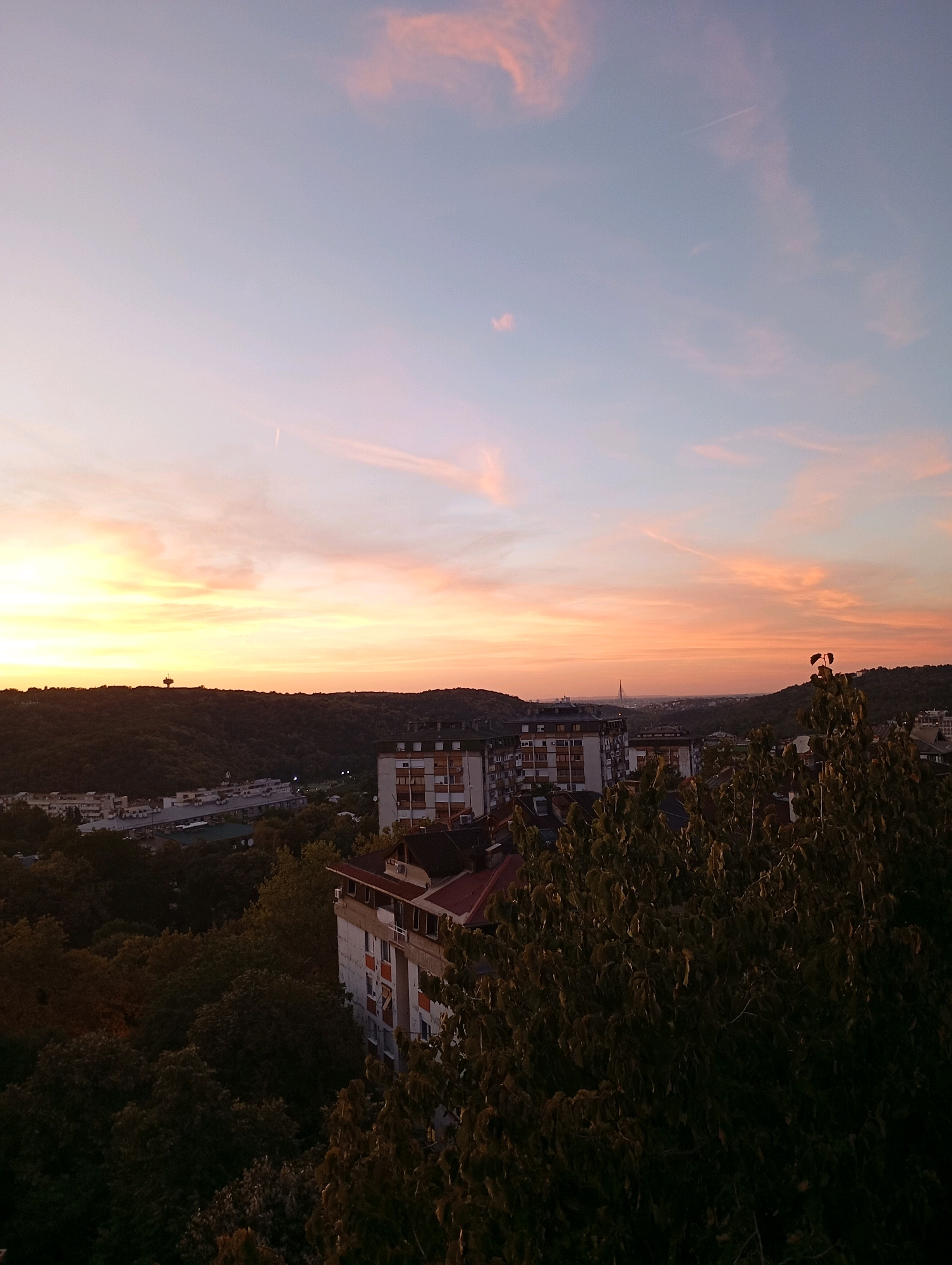 The image depicts a panoramic view of Belgrade, Serbia, at dusk. The sky is a canvas of soft pink and orange hues, with wisps of clouds streaking across it. In the distance, the silhouette of the Avala Tower is faintly visible against the horizon, a prominent landmark on the outskirts of the city. The foreground is dominated by lush green trees and foliage, with the tops of apartment buildings peeking through. The buildings appear to be typical of Belgrade's residential areas, with concrete facades and balconies. The scene suggests a peaceful evening in the city, with the urban landscape blending harmoniously with the natural environment. There are no people or animals visible in the image, and no specific actions or interactions are taking place. The overall atmosphere is tranquil and serene, capturing the beauty of a sunset over an urban setting.
