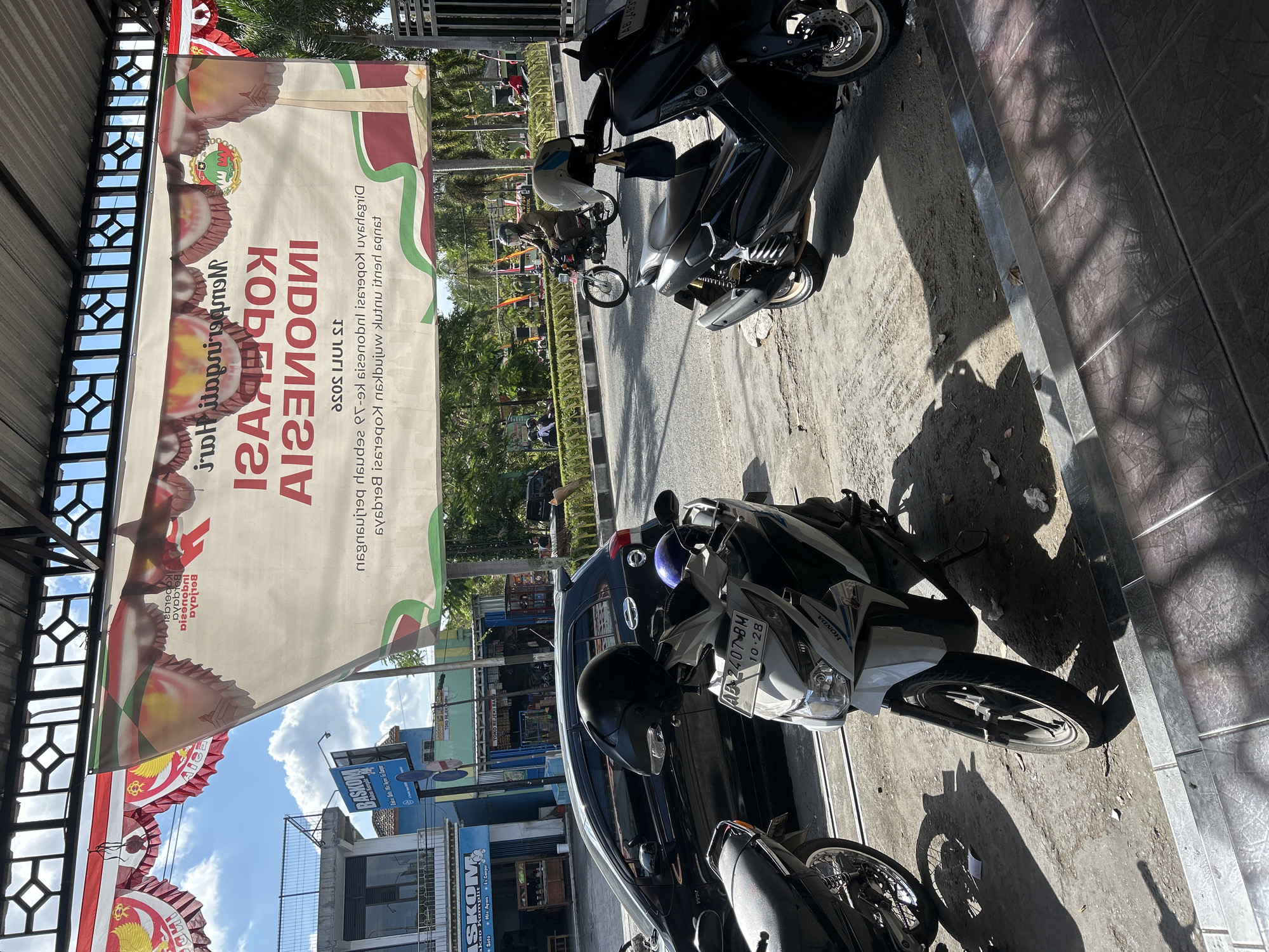 This image captures a street scene in Wonosari, Indonesia, dominated by a large banner celebrating the "Hari Koperasi Indonesia" (Indonesian Cooperative Day). The banner, partially obscuring the sky and buildings, is white with red and green accents and features text and imagery related to the event. In the foreground, several motorcycles are parked on a sidewalk. A white Honda scooter with the license plate "AD 2407 BM" is prominently featured, along with a black maxi-scooter to its right. A black car is parked on the street behind the motorcycles. On the road, a person wearing a helmet and a brown jacket is riding a motorcycle, and other individuals can be seen in the background. The environment suggests a commercial area with buildings, trees, and a sunny sky with scattered clouds.