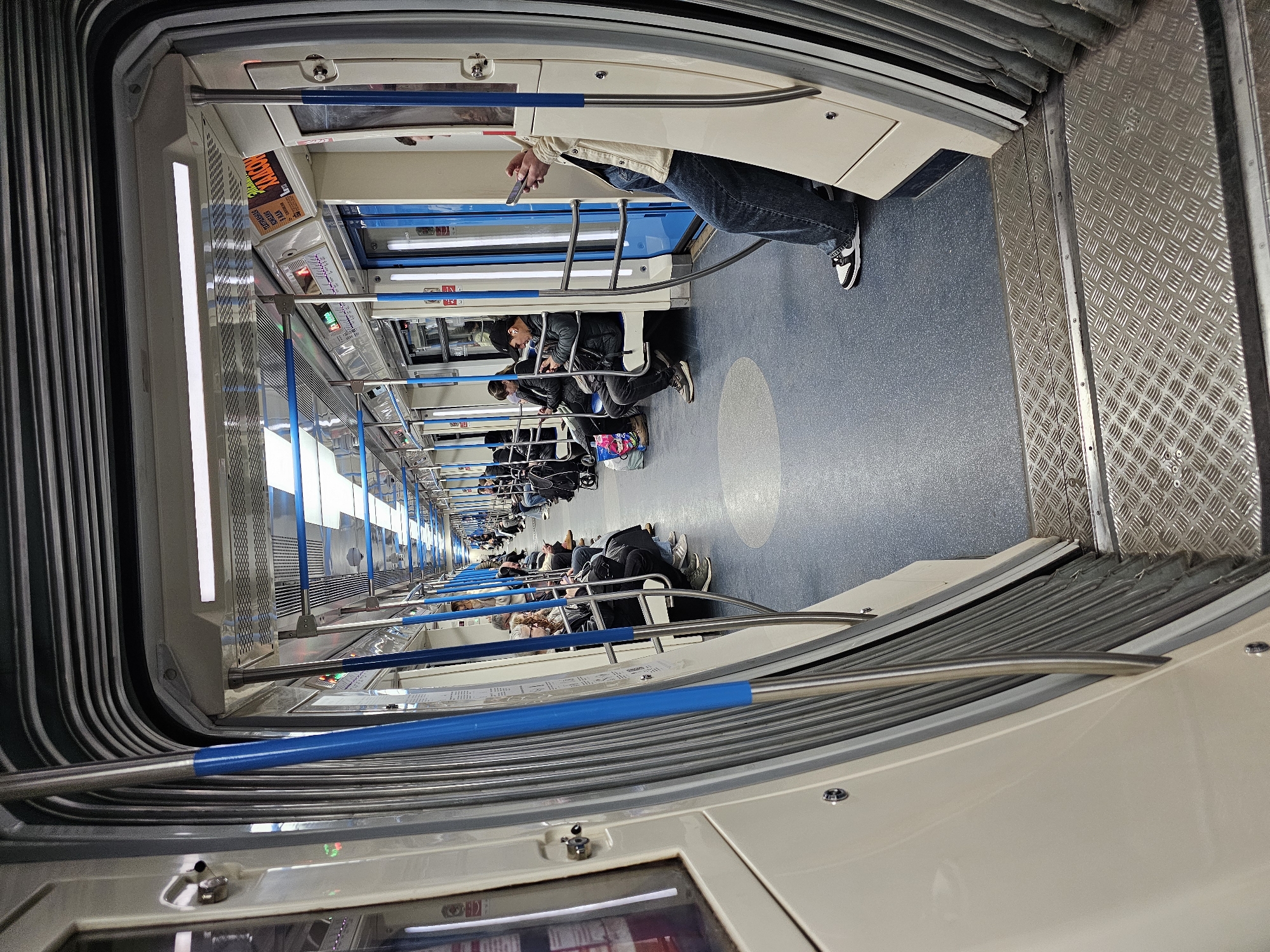 Inside a Moscow metro car, passengers are seated along the sides. The car's interior features blue accents. A person near the camera uses a phone. The floor is blue. A textured metal surface is visible near the edge.
