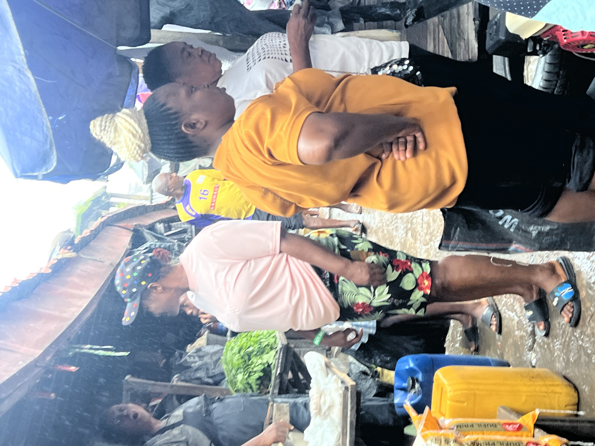 In Nkpor, Nigeria, a bustling market scene unfolds under a light rain. Several women are the primary subjects, with one in the foreground wearing an orange t-shirt and black shorts, her hands clasped behind her back. To her left, another woman in a pink t-shirt and floral shorts looks away, wearing a colorful baseball cap. Behind them, other individuals are partially visible. The environment is characterized by outdoor market stalls with corrugated roofing, suggesting a covered but open-air setting. Various objects are scattered around, including large yellow and blue plastic containers, bags, and crates, indicating the sale and storage of goods. The ground is wet and appears to be muddy, consistent with the rainfall. The overall atmosphere is that of a typical day in a vibrant Nigerian market.