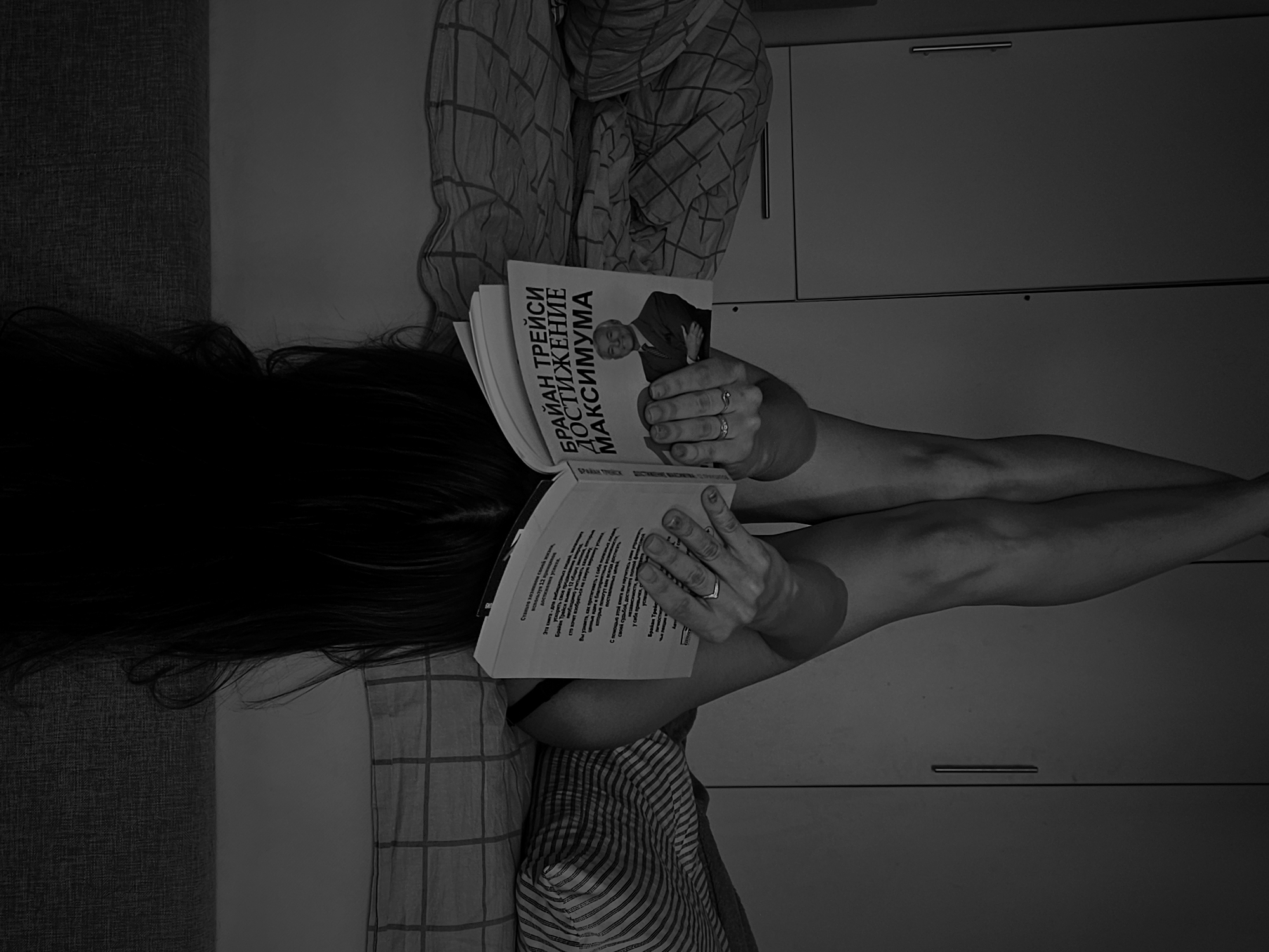 A person is lying on a sofa in a room in Chaikovsky, Russia, reading a book titled "Brian Tracy. Maximum Achievement" in Russian. The person has long dark hair and is wearing a striped shirt. They are holding the book open with both hands and have rings on their fingers. The sofa has a textured fabric. Behind the person is a wall or cabinet with a horizontal handle. The image is in black and white.