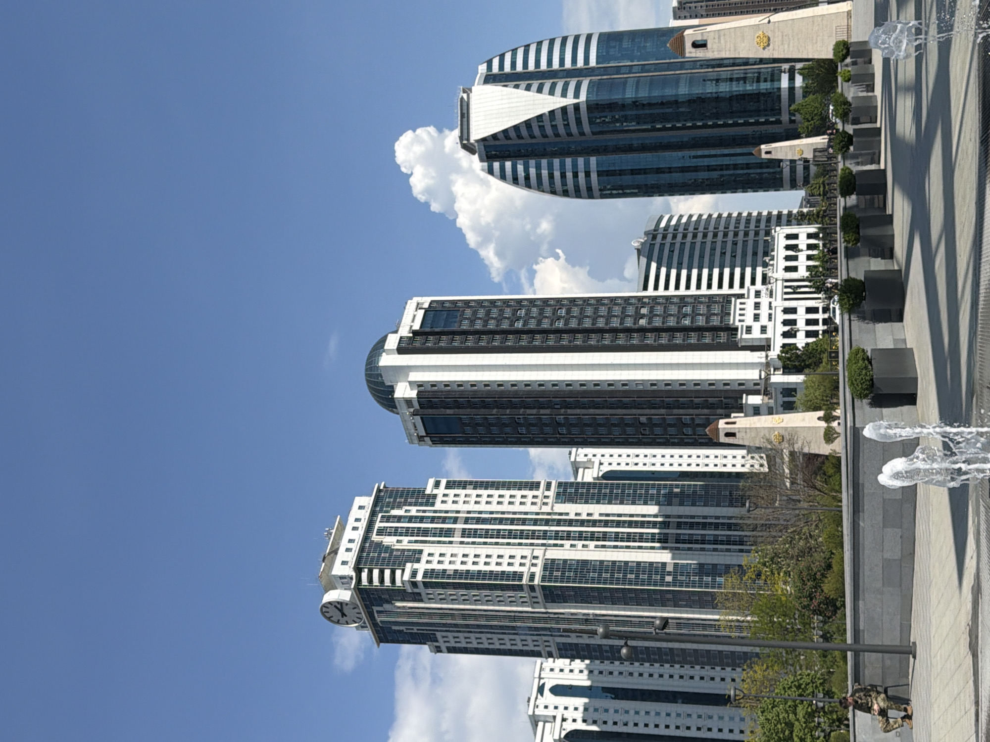 The image depicts a street scene in Grozny, Russia, featuring several tall, modern buildings against a partly cloudy, bright blue sky. The buildings are of varying heights and designs, with some featuring clock towers and others with curved glass facades. In the foreground, there's a paved pedestrian area with a fountain spouting water. A person in camouflage clothing is visible near a lamppost on the street, possibly a security guard or military personnel. There are also some small trees and bushes planted in the vicinity. The overall atmosphere is urban and modern.