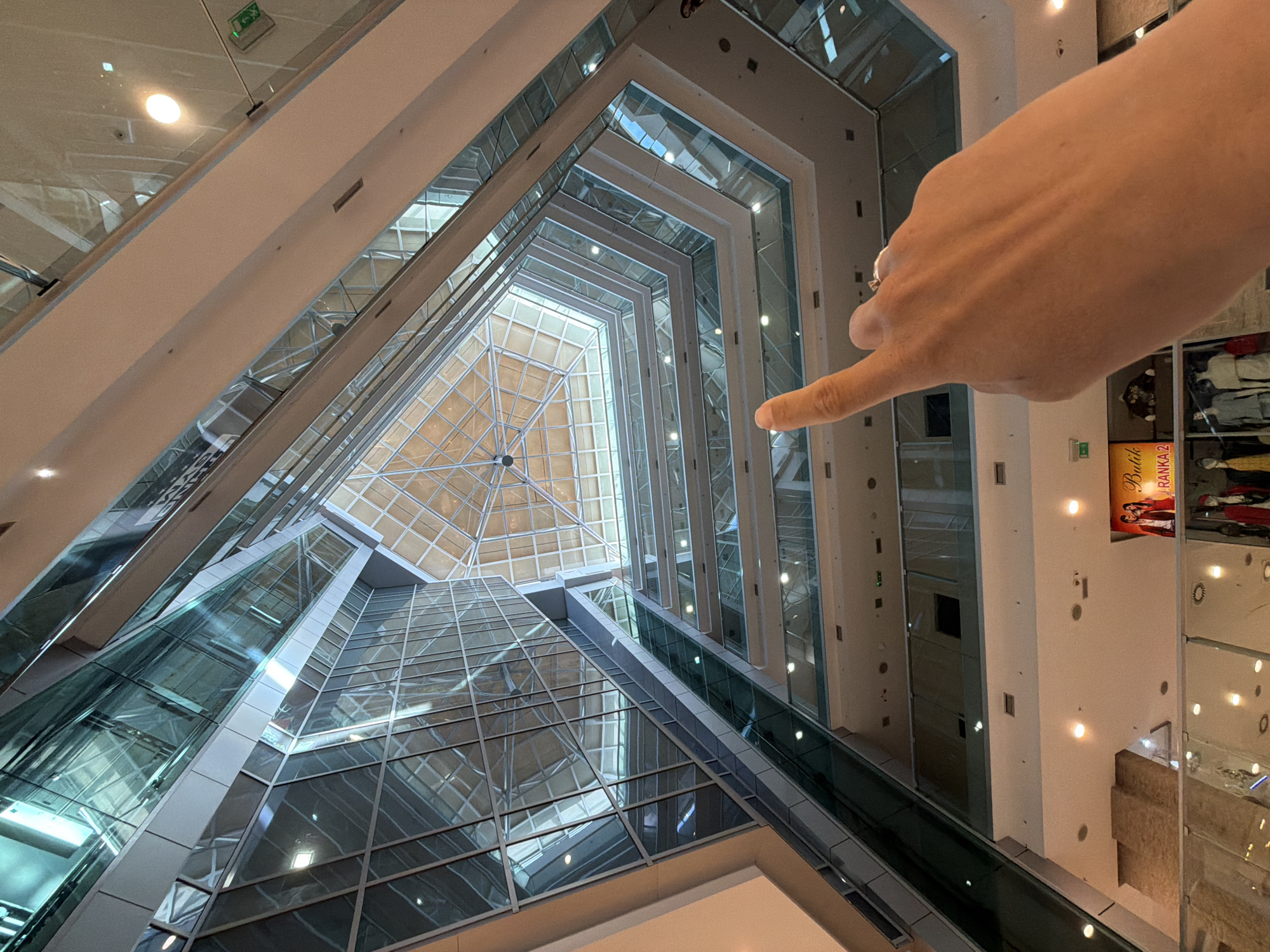 The image captures the interior of a modern, multi-story building, likely a shopping mall or hotel atrium, located in Budva, Montenegro. The perspective is from a lower floor, looking upwards into a vast central void, emphasizing the height and angular architecture.
In the foreground, on the right side, a human hand, possibly female, with a ring on the ring finger, extends into the frame, pointing towards the upper-middle section of the atrium. The building features extensive use of glass, creating numerous reflections of the interior structure, lighting, and distant elements. Multiple levels are visible, characterized by white walls, glass railings, and geometric patterns formed by the building's framework.
A prominent feature is the large, multi-faceted glass skylight at the very top of the atrium, allowing natural light to filter in. Escalators with glass sides are seen traversing the space, connecting different floors. Recessed ceiling lights illuminate various sections of the building.
Notable details include an emergency exit sign with a green running figure visible in the upper left section. On a wall in the mid-ground to the right, a poster or advertisement is visible, displaying images of people and text, including "Buš" and "FRANKA 2". The overall impression is one of a spacious, well-lit, and architecturally striking contemporary interior.