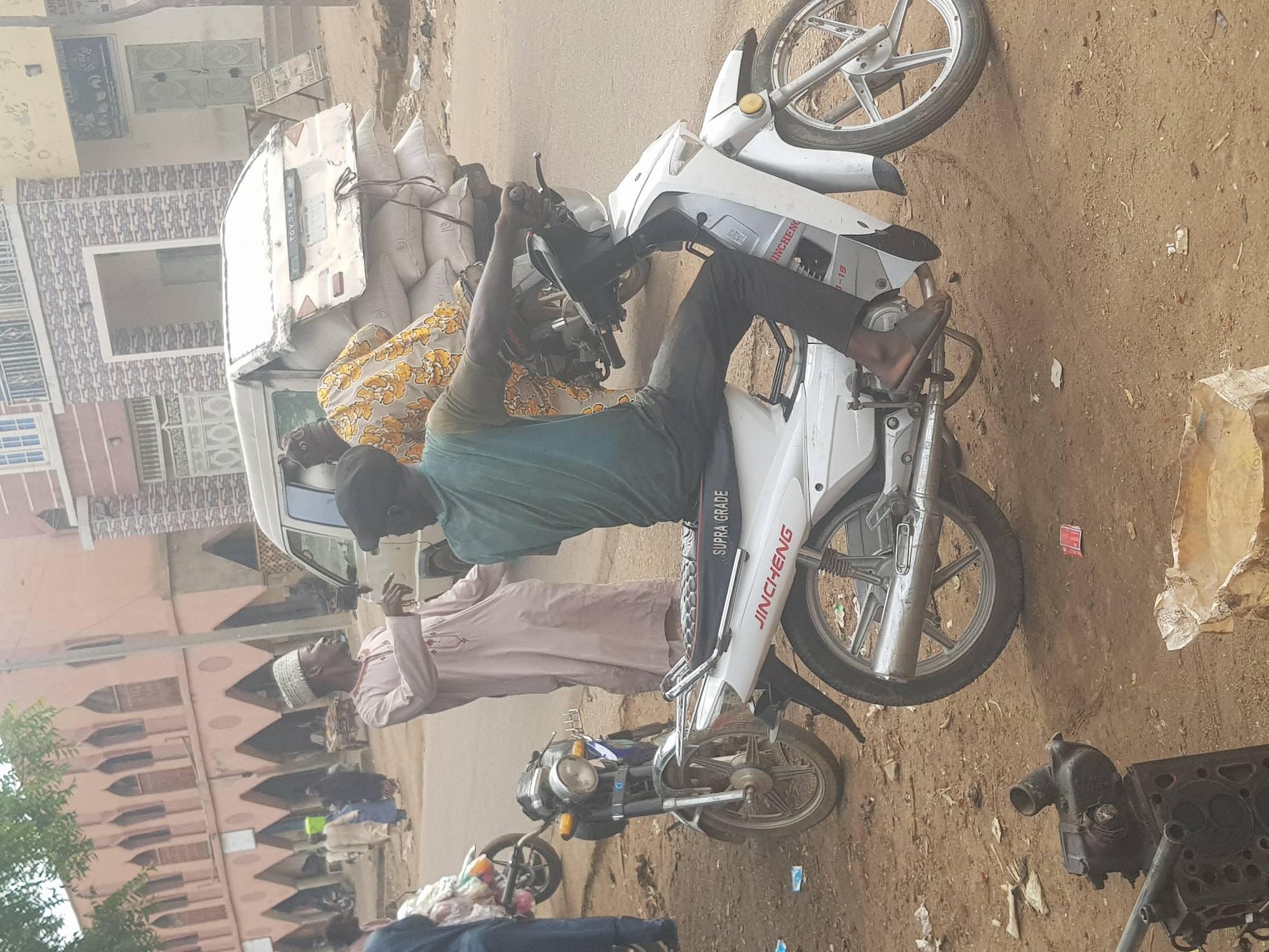 The image shows a street scene in Kufena, Nigeria. A man in a green shirt is seated on a white Jincheng Supra Grade motorcycle. Another man in a light pink robe and a white cap stands nearby. In the background, a white truck loaded with sacks is visible. Several other motorcycles are parked along the street. Engine parts are seen on the ground near the motorcycle. The environment is dusty and appears to be a commercial or residential area with buildings lining the street.