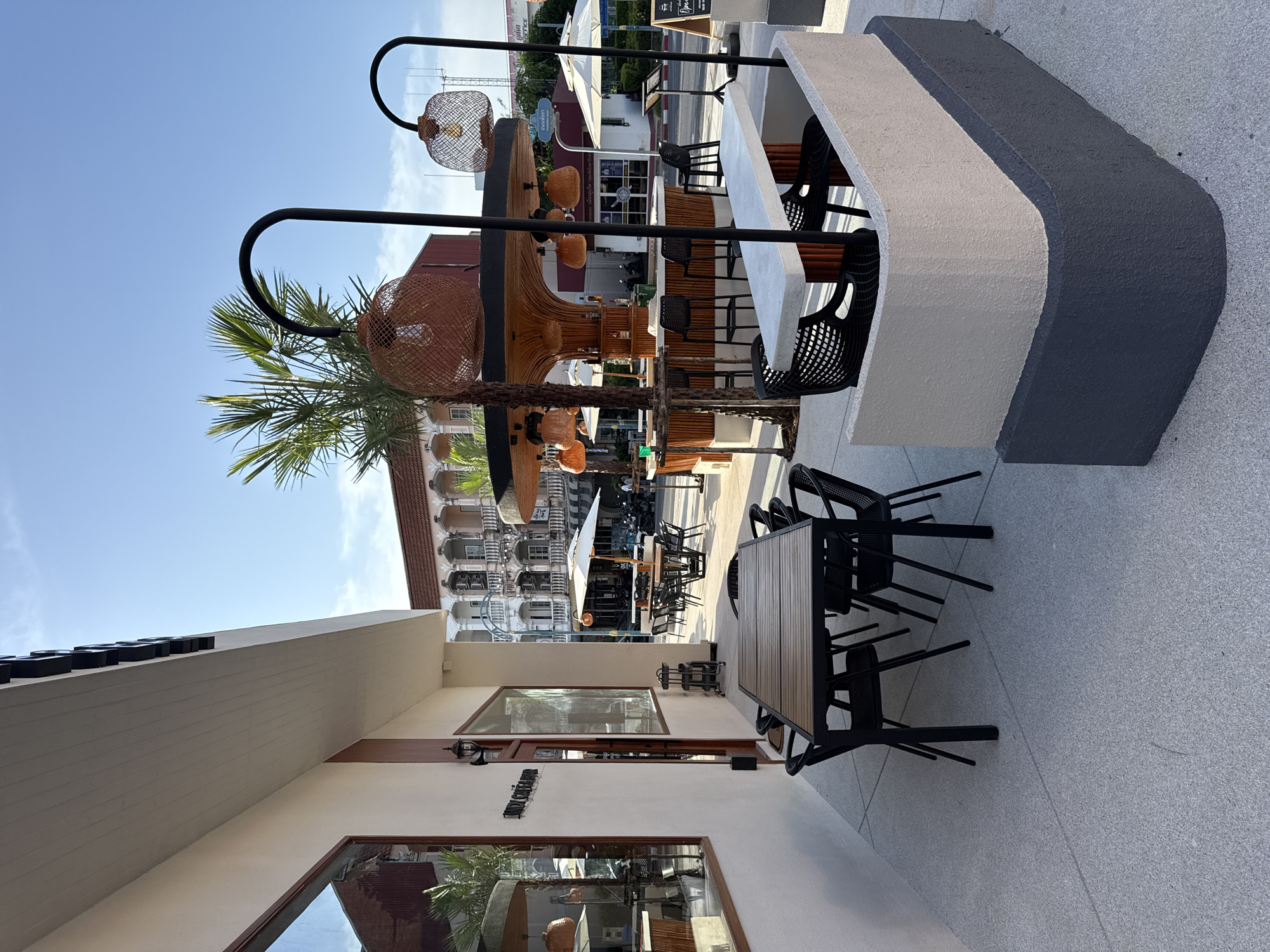 The image depicts an outdoor seating area of a cafe or restaurant, likely located in Phuket City Municipality, Thailand. The foreground features a square table with black chairs, set on a light-colored, textured pavement. To the right, a curved, two-tiered planter box, painted in beige and dark gray, separates the seating area from a sidewalk and street.
Further into the scene, a circular bar with a thatched roof and wooden accents is visible. Several bar stools are arranged around it, and bottles of alcohol are displayed on shelves behind the counter. Hanging light fixtures with woven shades are suspended from black poles. Palm trees are interspersed throughout the outdoor area, adding to the tropical ambiance.
In the background, traditional multi-story buildings with ornate architectural details line a street. Umbrellas and more tables and chairs suggest additional outdoor dining spaces. The sky is clear and blue, indicating fair weather. There are no people clearly visible in the immediate foreground or midground, though a few figures can be seen in the distance near the street. The overall impression is of a vibrant and well-maintained commercial establishment in a tropical urban setting.