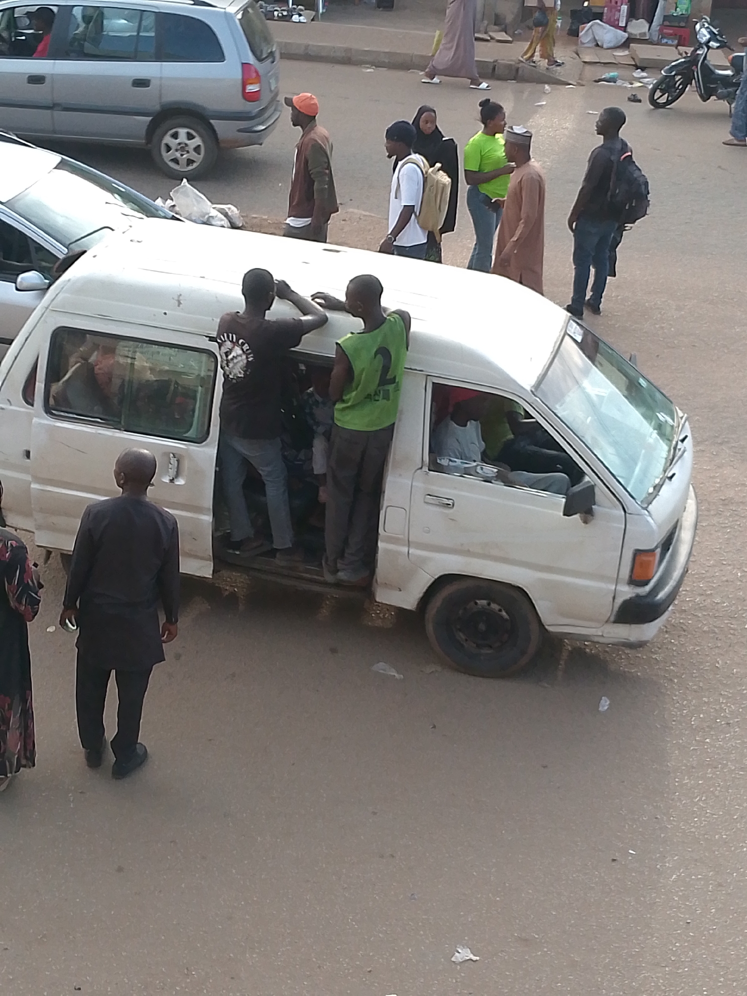The image depicts a lively street scene in Bukuru, Nigeria, during daylight hours, likely mid-morning or afternoon, characterized by clear and dry weather. The central element is a white minivan, appearing to be a public passenger vehicle, with its side sliding door open.
Two men are positioned at the open door: one in a black t-shirt with an illegible white design on the back and jeans, and another in a green sleeveless top bearing the clear white number "2" on his back, wearing dark trousers. They appear to be either embarking, disembarking, or handling items. Several passengers are visible inside the minivan, including a driver.
In the foreground, a man in a black long-sleeved tunic and trousers walks away from the minivan. In the background, a silver station wagon, resembling an Opel/Vauxhall Zafira, is visible. Near it, a man in an orange cap and a brown jacket stands. Further into the background, a group of people is seen interacting on the paved street: a woman in a bright green top, a man in a brown traditional garment, a woman in a black headscarf holding a light-colored jerrycan, and a man carrying a black backpack. A motor scooter is parked on the right side of the street, and other pedestrians are visible in the distance. The road surface shows some scattered litter, including plastic bags. The overall scene conveys typical daily activities in an urban or semi-urban environment.