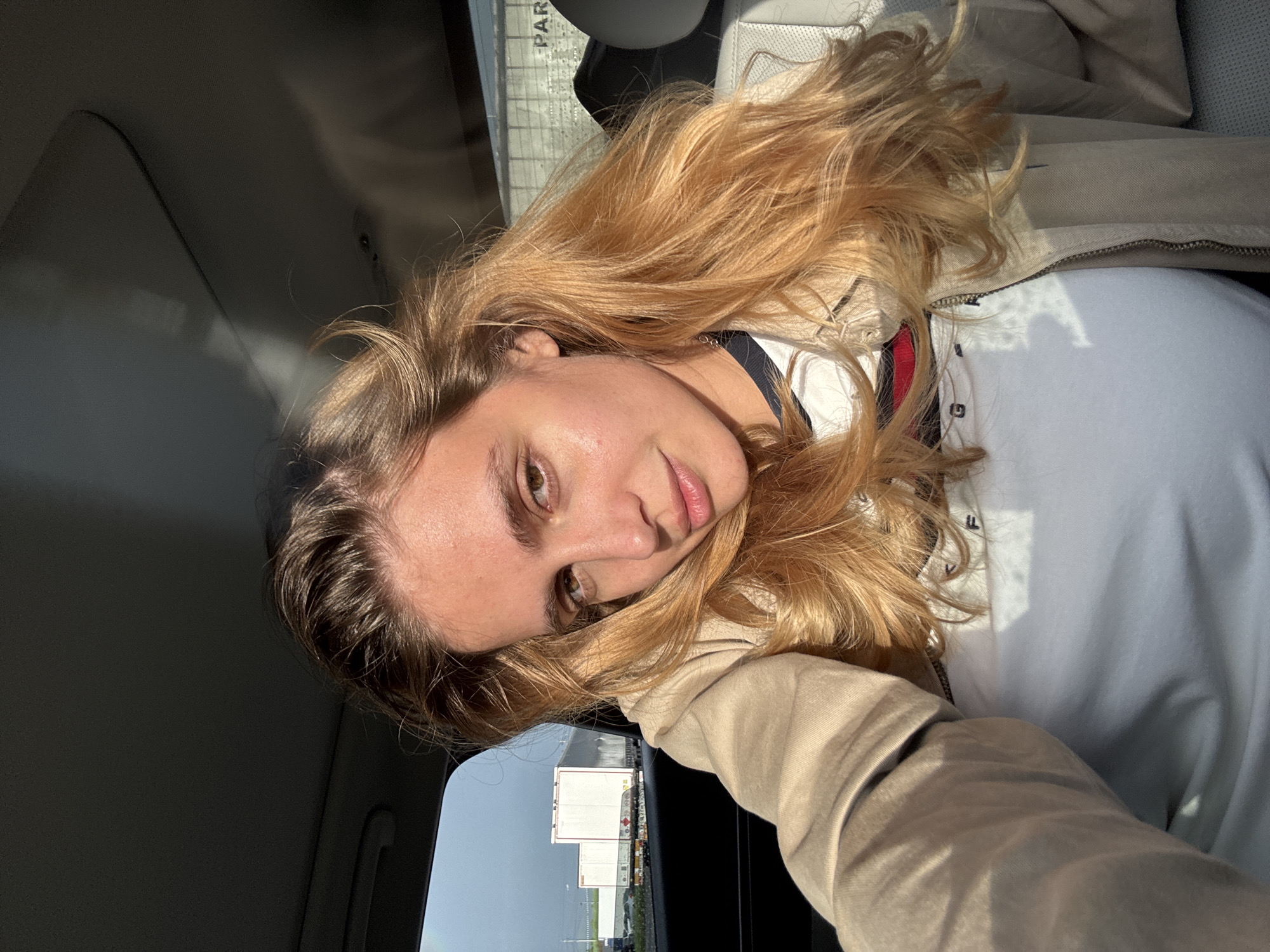 A woman with long blonde hair is taking a selfie inside a car. The background visible through the car window shows a building and the streetscape of what appears to be an industrial area. The sign "PARKING" is visible in the top left corner, likely within the parking lot of the building seen. There is no definitive evidence in the image to confirm the location as Bagratashen, Armenia.