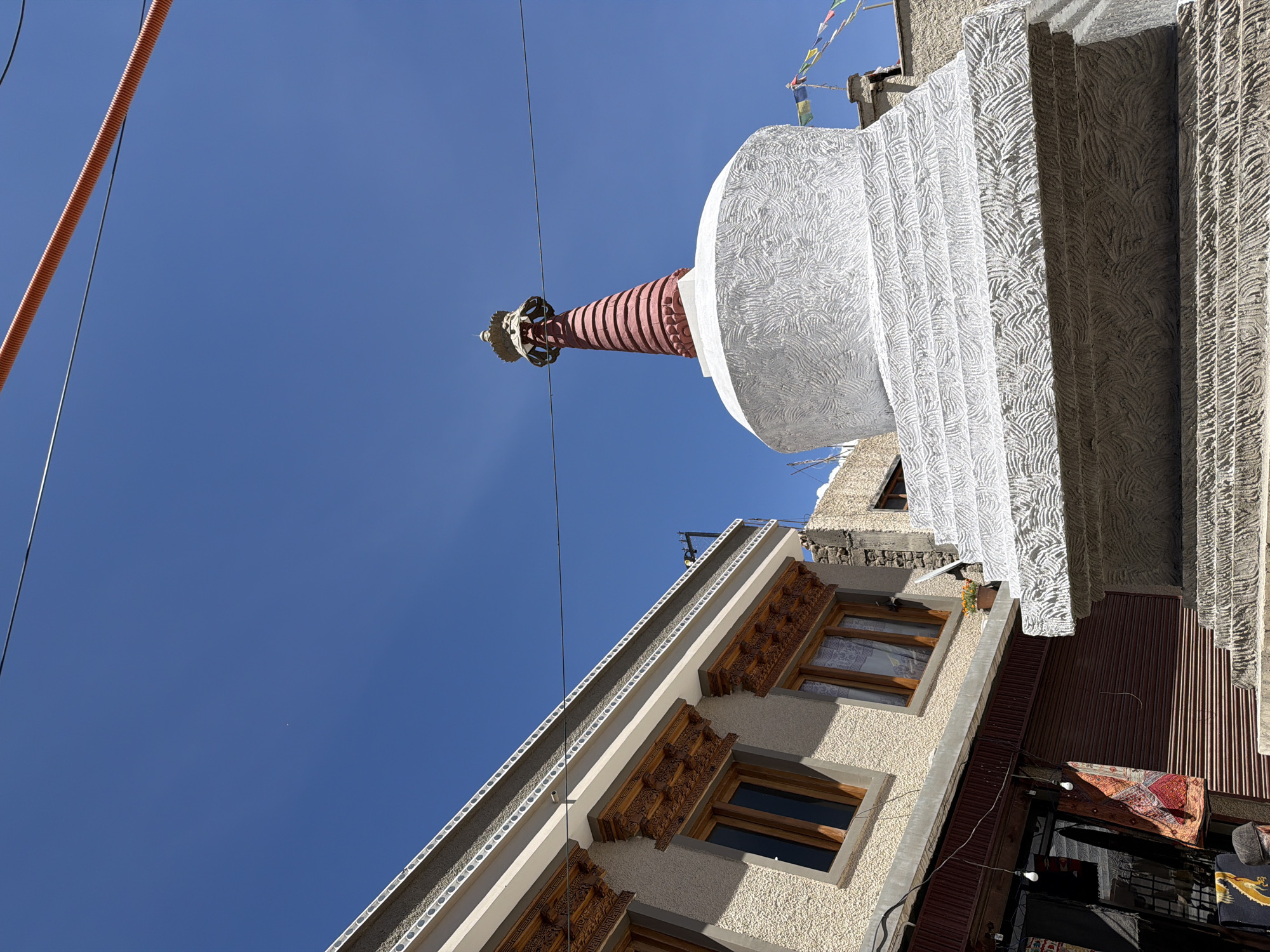The image, taken in Leh, India, depicts a clear blue sky above a stupa and a building. The stupa, prominently featured, is predominantly white with a textured surface on its base and a red, conical spire topped with a golden finial. Prayer flags flutter on a structure behind the stupa. To the left, a building with light-colored textured walls and wooden-framed windows is visible. The windows feature intricate wooden carvings above them. Below the building, a shop with closed metal shutters and displayed textiles can be seen, with a glimpse of a person wearing a cap. The environment suggests an outdoor urban setting with a strong cultural and religious presence.