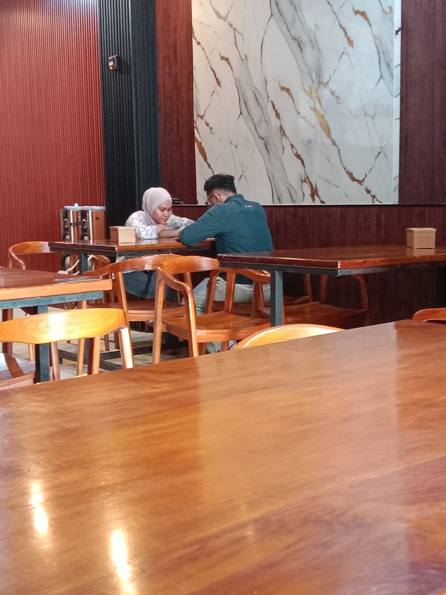 The image depicts two people seated at a table in a restaurant. A woman wearing a light pink hijab and a patterned blouse is sitting across from a man with dark hair and glasses, wearing a blue shirt. They are both leaning on the table. The restaurant has wooden tables and chairs. The background includes a red wall with a black ribbed section, and a marble-patterned decorative panel. The location is Pandean, Indonesia.