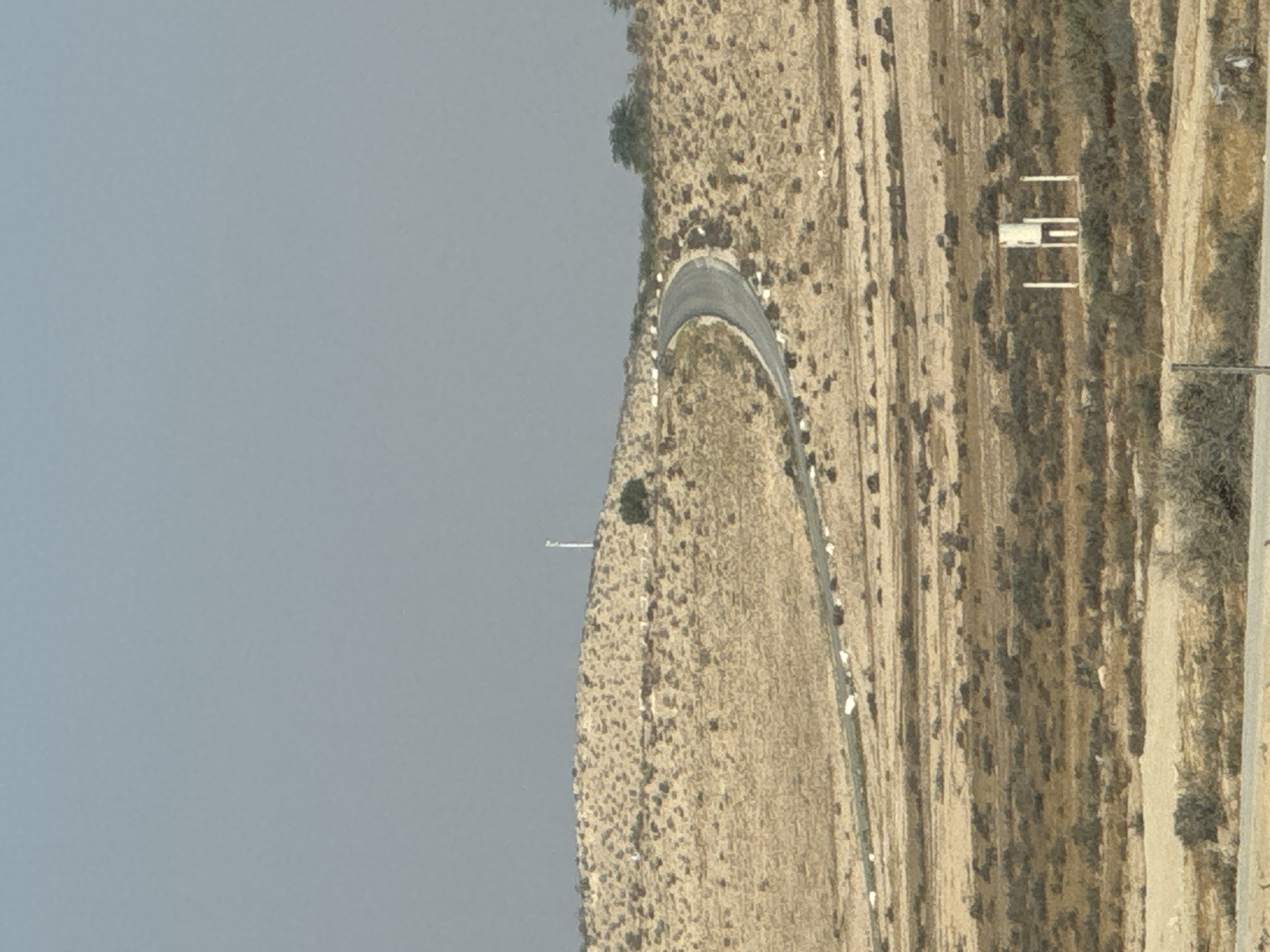 The image captures a desolate, arid landscape within the Ramat Negev Regional Council, Israel. A paved, winding road snakes its way across a tan, rocky terrain dotted with sparse, dry vegetation. Along the roadside, white markers are visible, indicating the edge of the pavement. In the distance, atop a hill, a tall, slender white structure with a rectangular top stands against a pale, hazy sky. The environment suggests a remote, possibly desert-like region.