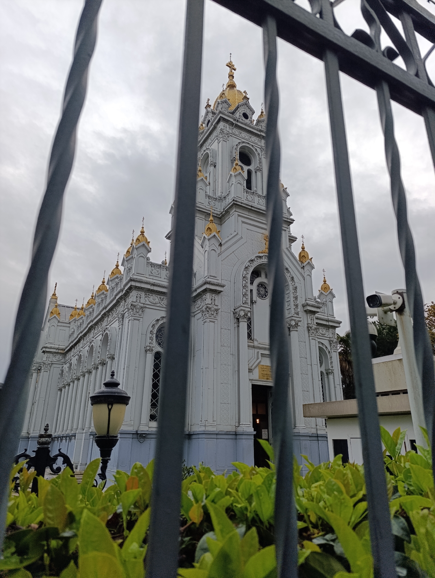 The image depicts the Bulgarian St Stephen Church, also known as the Iron Church, in Istanbul, Turkey, viewed through a metal fence. The church is white with golden domes and ornate architectural details. A lamppost stands in front of the church, partially obscured by green bushes. To the right of the church, a white building with a security camera is visible. The sky is overcast.