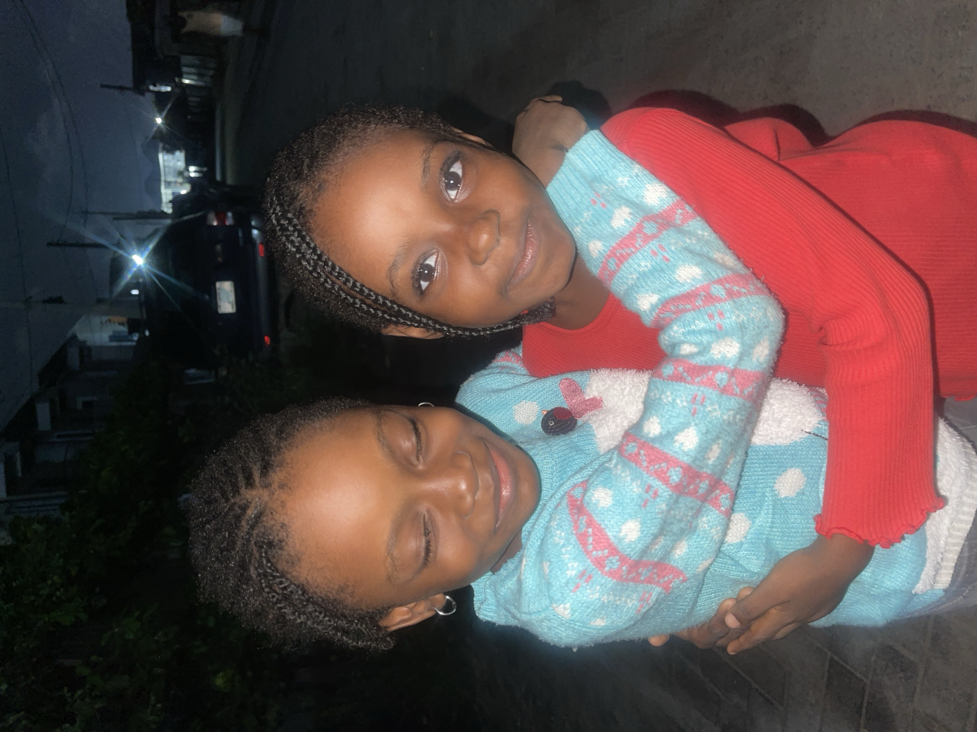 Two young girls are embracing on a street at night. The girl on the left is wearing a light blue sweater with white and pink patterns and her eyes are closed as she smiles. The girl on the right is wearing a red long-sleeved shirt, and her eyes are open as she looks at the camera. They are standing in what appears to be a residential street in Ado, Nigeria, with buildings and a parked vehicle visible in the background. The lighting suggests it is nighttime.