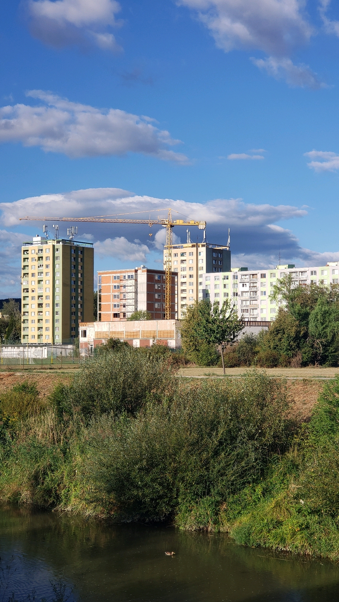 A crane is visible over a cluster of apartment buildings in Prešov, Slovakia. The crane is yellow, and it is positioned to the left of the frame, with its arm extending horizontally across the sky. The apartment buildings vary in height and color, with some being tall and slender, while others are shorter and wider. One building in the foreground is green and yellow, and it has many windows. Another building behind it is orange and appears to be under construction. The sky is blue with scattered white clouds. In front of the buildings is a body of water, possibly a river, with trees and bushes on its banks. A duck is swimming in the water. The overall scene suggests a city landscape with ongoing development.