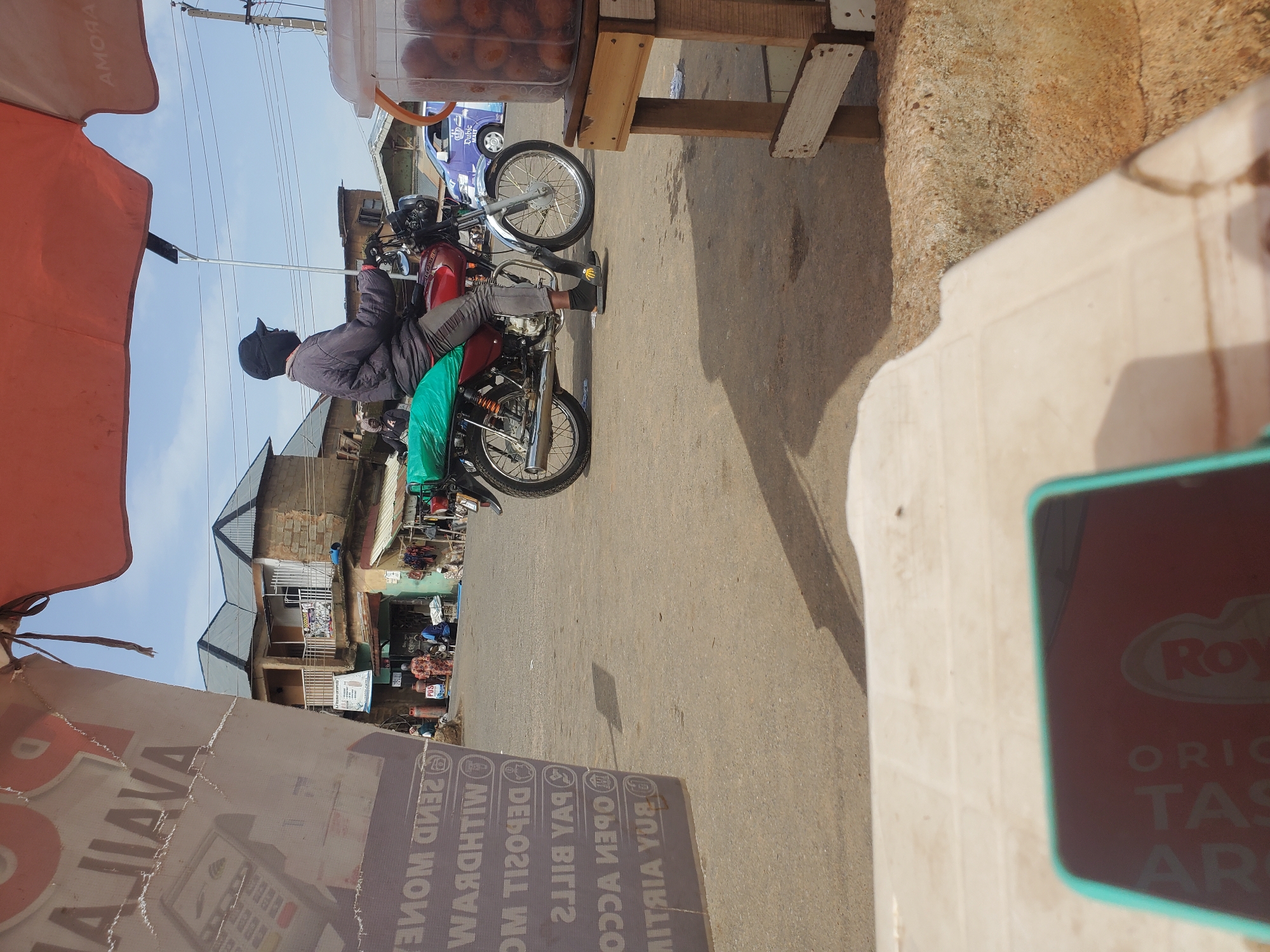 The image depicts a street scene in Gwong, Nigeria, characterized by a bustling marketplace environment under a bright, possibly midday sun. In the foreground, a partially visible table and a smartphone with a turquoise case suggest a seated observer. Dominating the mid-ground is a man wearing a dark jacket, dark pants, black socks, and black sandals, astride a red motorcycle. The motorcycle has a green tarp strapped to its rear. The man is wearing a black face mask and a black baseball cap, obscuring his facial features. He is looking to his left, and his hands are on the handlebars.
Behind the motorcycle, a variety of buildings line the street. Some appear to be shops or residential structures, with multiple stories and balconies. A prominent advertisement, partially torn, features text that appears to be in English, with legible phrases such as "SEND MONEY" and "WITHDRAW." Other signs are visible, including one for "POS" (Point of Sale). Several other people are present in the background, some walking and others seemingly gathered near the shops. One person is wearing a vibrantly patterned traditional outfit. A purple vehicle is also visible on the road.
To the right of the motorcycle, a wooden stand holds a clear plastic container filled with what appear to be small, spherical food items, possibly fritters or dumplings. The overall environment suggests a lively urban setting where commerce and daily life intersect. The pavement is paved, with some patches of dirt or uneven surfaces. Power lines are visible overhead.