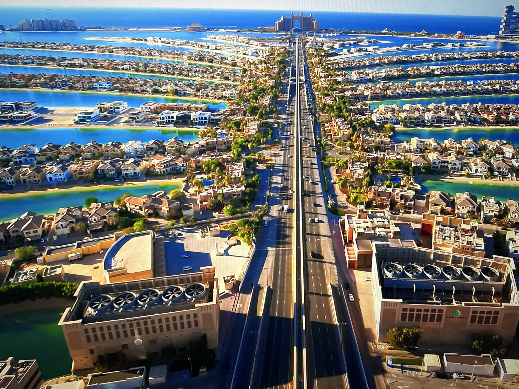The image displays an aerial view of a developed coastal area, characterized by artificial islands or peninsulas extending into a body of water. The structures on these landmasses appear to be residential, with many individual houses featuring distinct architectural styles, often with light-colored facades and tiled roofs. These residential areas are interspersed with lush green vegetation and bordered by canals or waterways.
A prominent feature is a multi-lane highway that runs centrally through the scene, connecting the developed area to a larger landmass in the distance. The highway is populated with vehicles, indicating ongoing traffic. Along the sides of the highway, there are several large, industrial-looking buildings, one of which prominently features a sign with the word "EMPOWER" and is topped with multiple large circular ventilation units.
In the background, a large, distinct hotel or resort complex is visible, situated on a prominent man-made island or structure. The water is a vibrant blue, and the sky is clear and bright, suggesting a sunny day. The overall impression is one of a highly planned and developed resort or residential community.
The scene is not located in Burgas, Bulgaria.