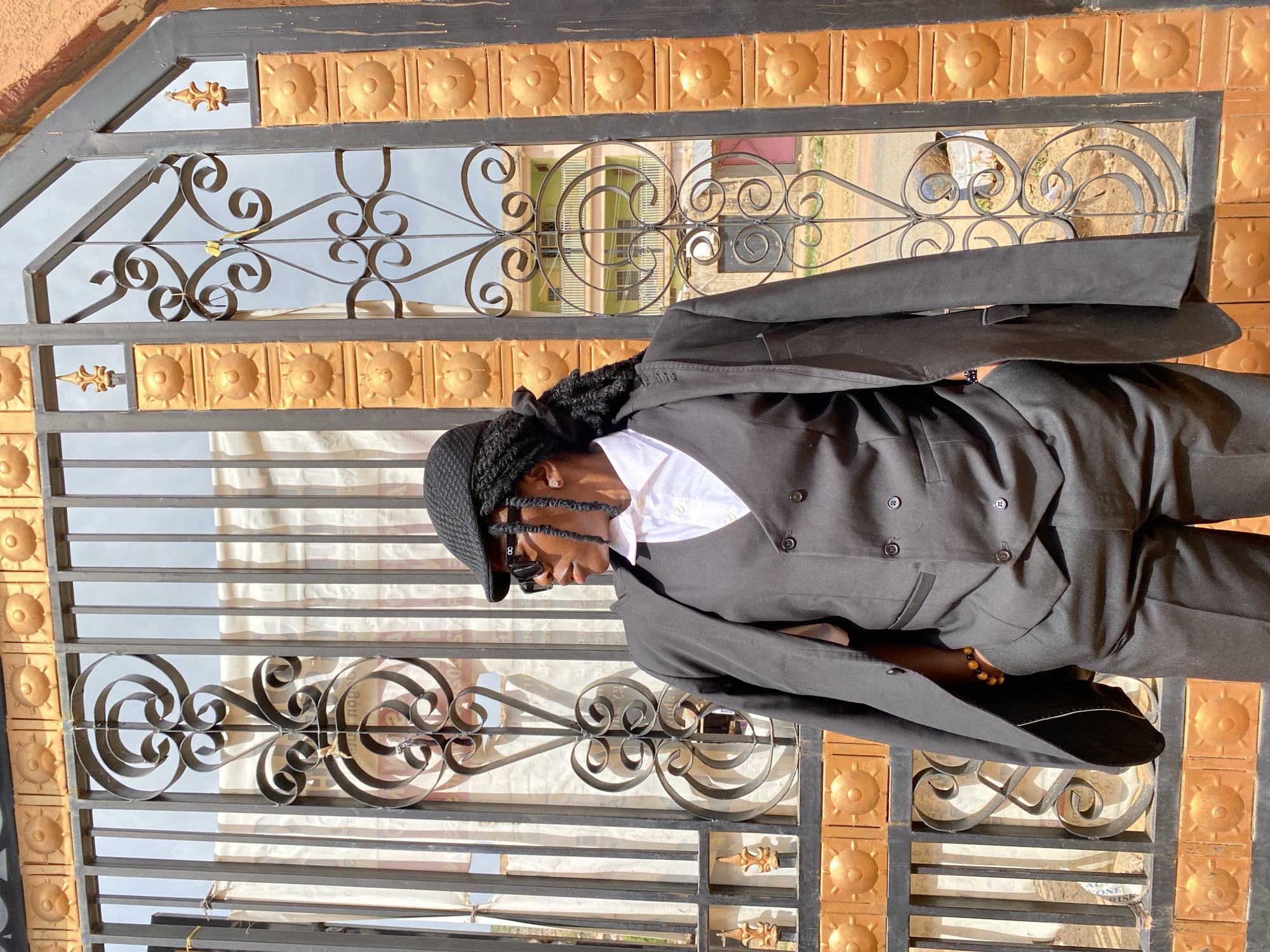 A person wearing a black suit, white shirt, black hat, and sunglasses stands in front of a black and gold metal gate in Gwong, Nigeria. The gate has decorative wrought iron details. A building can be seen through the gate.