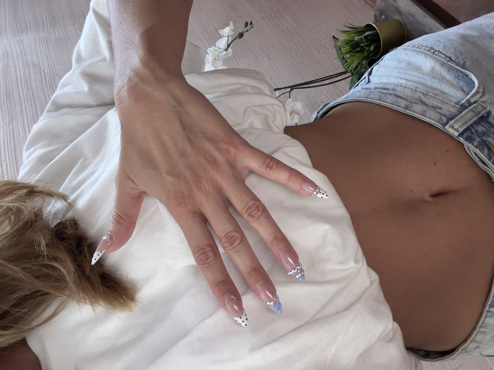 The image shows a close-up of a person lying down on a white surface, possibly a bed or floor. The person is wearing light blue denim shorts, and their abdomen is visible. A hand with long, decorated fingernails rests on the person's torso. There are also white flowers and a small potted plant visible in the background. The image was taken in North Kuta, Indonesia.
