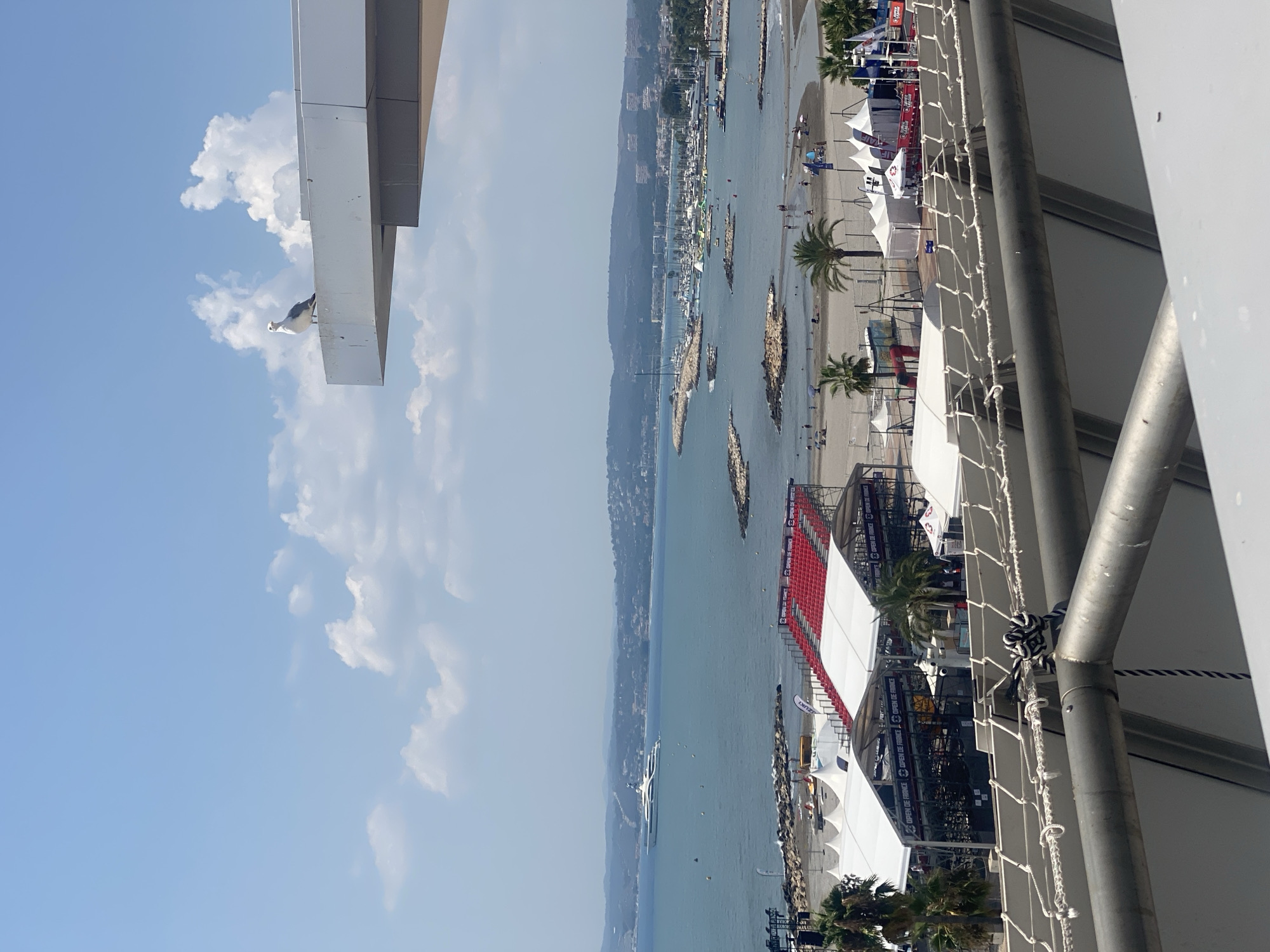 An elevated, wide shot shows a view of the coast of Saint-Laurent-du-Var, France, on a bright, sunny day. In the foreground, a white railing and metal structure are visible. A seagull is perched on the edge of a white overhang in the middle ground, to the right. Beyond this, a beach area is set up with white tents, a stage, and red stadium seating. The sea stretches out to the horizon, dotted with several boats, including a large white yacht. The coastline is lined with buildings and hills, and a clear blue sky with scattered white clouds completes the scene.