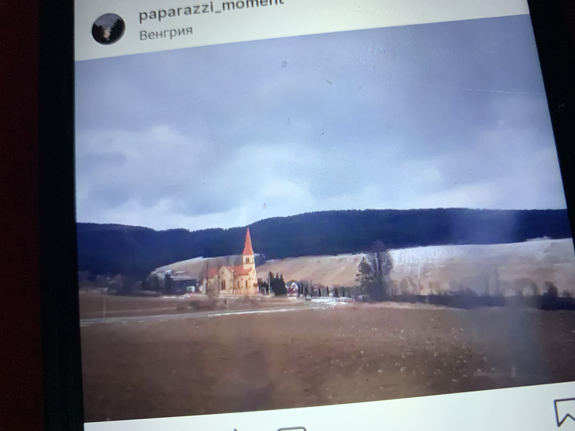 The image shows a rural landscape with a body of water in the foreground and a building with a red roof situated on a hill in the background. The sky is overcast. Text overlays on the right side of the image include "paparazzi_moment" and "Венгрия" (Hungary in Russian). There is no visual information to confirm the location as Lymanka, Ukraine.