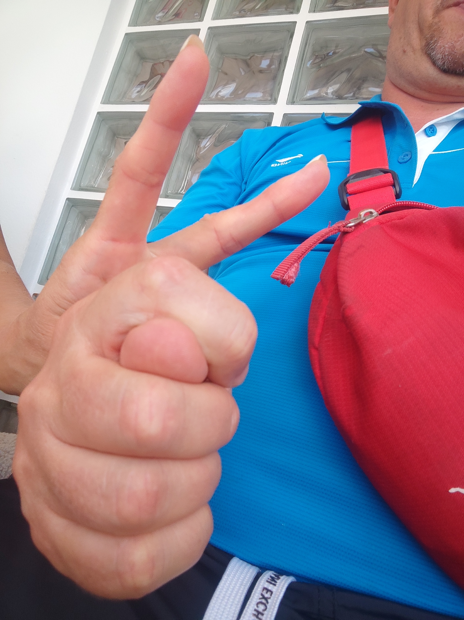 A close-up, eye-level shot features a person's left hand, held up in a peace sign gesture. The hand is in focus, with visible skin texture and fingernails. Behind the hand, a person wearing a bright blue polo shirt with a white collar and a red crossbody bag is partially visible. The bag has a silver zipper and a red strap. The background consists of a wall with clear glass blocks, suggesting an indoor or covered outdoor setting, possibly in Bar, Montenegro. The lighting is bright, creating some highlights on the skin and fabric. The overall scene is casual and relaxed.