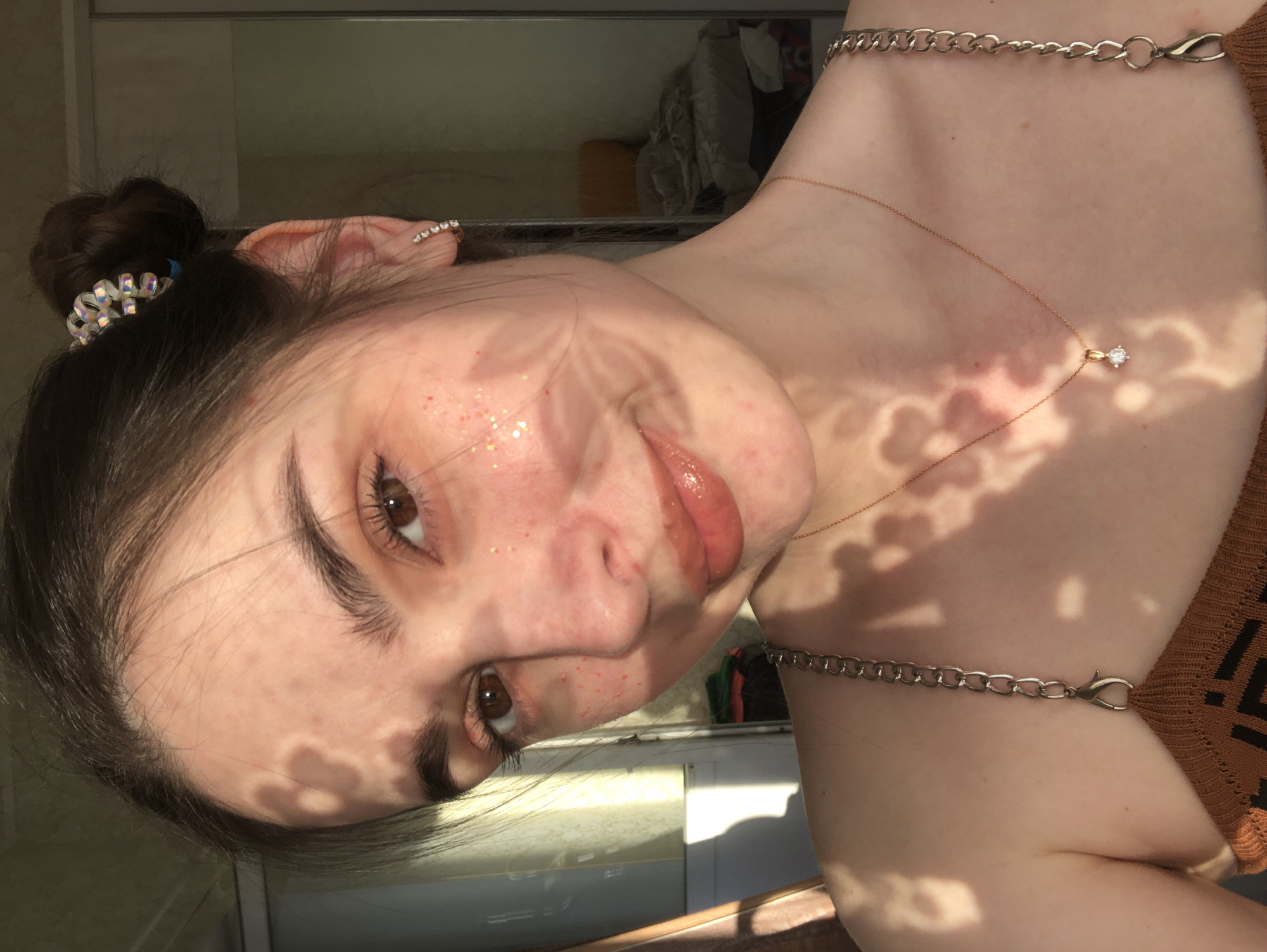 A medium shot shows a person's face and part of their upper body. The person has light skin, brown eyes, and dark hair pulled back in a bun with a sparkly hair tie. They are wearing a small, gold necklace with a diamond pendant and a silver chain across their shoulder. Sunlight filters through leaves, casting shadows on their face and upper chest. There is glitter or small, red dots on the face. The location is unknown, and there is no discernible connection to the city of Noviy, Russia.