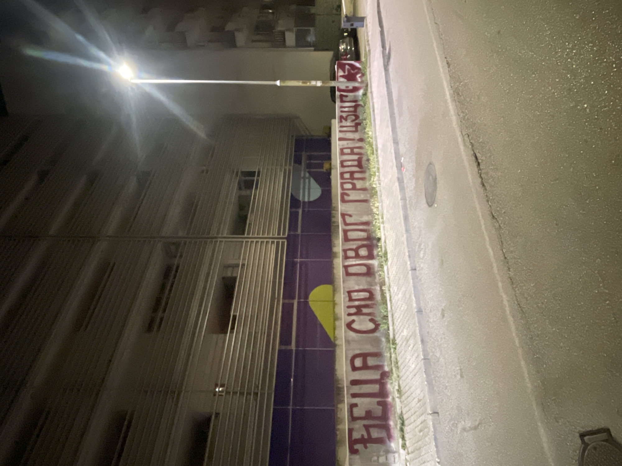 An outdoor night scene depicts a street in the city of Unknown, Unknown, illuminated by a single bright streetlamp on the right. A multi-story building occupies the left background, featuring a facade of horizontal light-colored slats above a wide purple band. On the purple band, a yellow, irregular semi-circular shape is spray-painted. Below this, a concrete wall runs horizontally across the image, covered in red Cyrillic graffiti: "ЂЕЦА СМО ОВОГ ГРАДА! ЦЗЦГ?". This translates to "WE ARE THE CHILDREN OF THIS CITY! CZCG?", with the final characters appearing as a "1" and possibly a "3" or an arrow. Sparse green foliage grows along the top edge of the concrete wall. In the foreground, a grey paver sidewalk with a curb abuts an asphalt street. A circular manhole cover is visible in the lower-left corner of the street. A dark-colored vehicle is partially visible parked on the street behind the lamppost, and a dark blue public trash receptacle stands on the sidewalk further right. The street lamp emits a strong glare, producing prominent light streaks across the upper right quadrant of the image.
