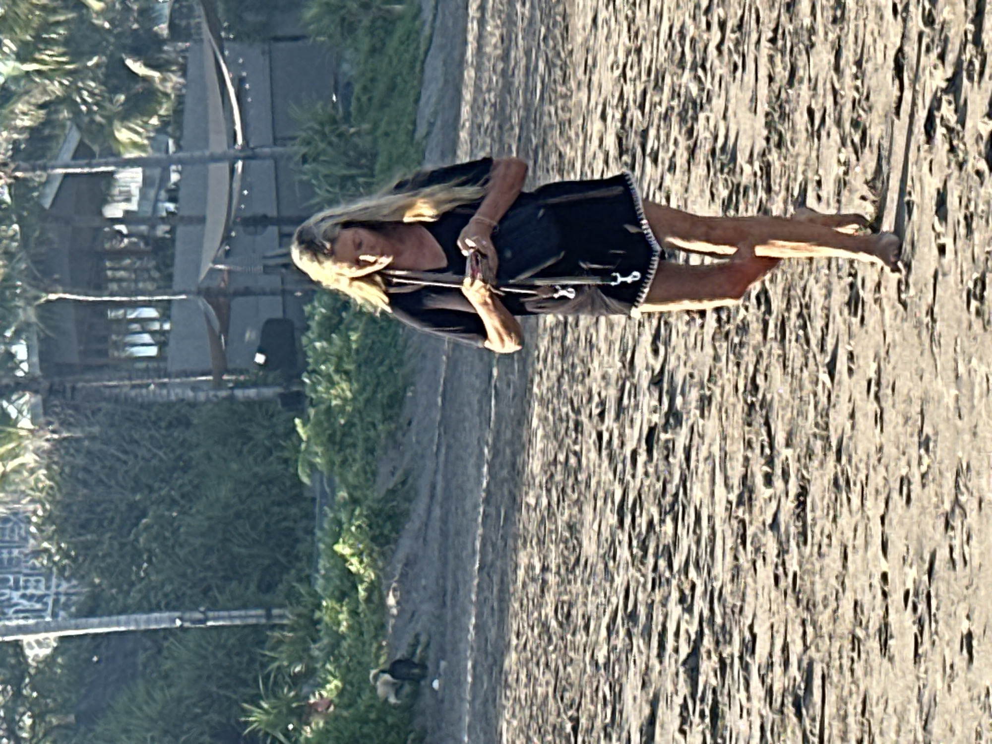 In North Kuta, Indonesia, a blonde woman walks on a sandy beach. She is wearing a black t-shirt dress and black shorts, carrying a black purse. Her hair is long and blonde. In the background, there are palm trees and a building with awnings. The sand is damp and textured, with footprints visible. The scene appears to be during daylight.