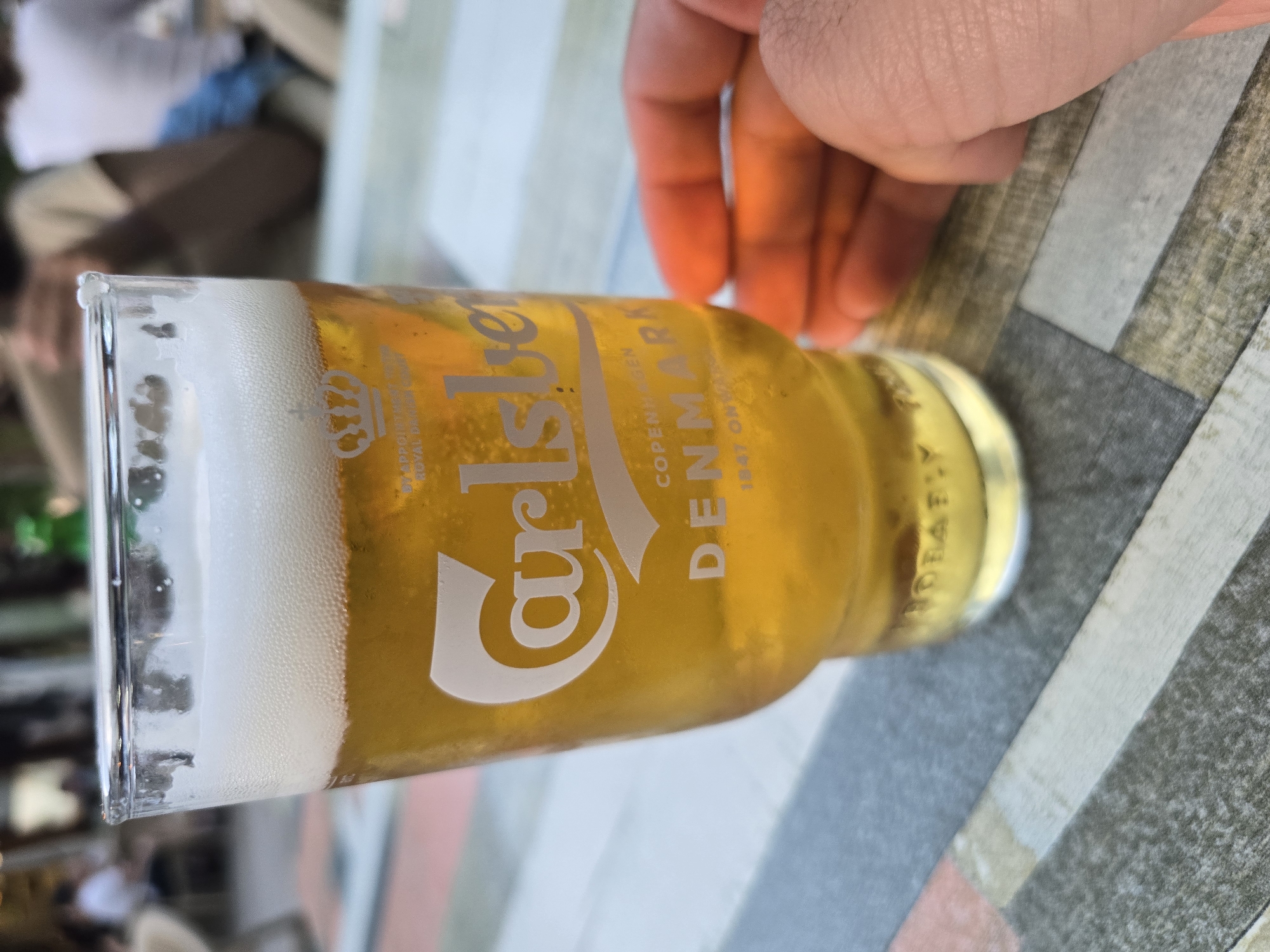 A close-up shot depicts a glass of Carlsberg beer, showing a substantial head of white foam at the top. The glass is adorned with the Carlsberg logo and the text "COPENHAGEN DENMARK" in white lettering. The liquid within the glass is golden and bubbly. The glass is situated on a tabletop that features a patterned surface with grey, white, and peach hues. A human hand is visible in the upper right corner of the frame, positioned near the glass. In the background, out-of-focus figures are present, suggesting an outdoor setting in an unknown city.