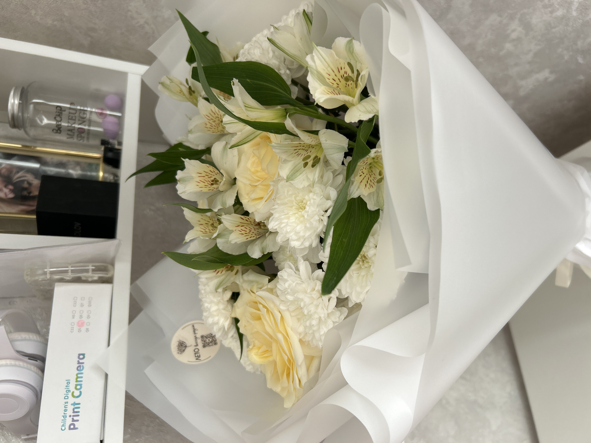 A white bouquet of flowers is presented in white paper. The bouquet is arranged with white alstroemeria, yellow roses, and white chrysanthemums, interspersed with green leaves. A small circular tag with Cyrillic text and a QR code is attached to the bouquet.
To the left of the bouquet, a white shelf unit is visible. On the shelf are several objects, including a clear plastic container labeled "Bee&Chip MAKEUP SPONGES" with purple sponges inside. Below this is a gold-framed picture. Further down, a black box is visible. Beneath the black box is a white box with text in both English and Russian. The English text reads "Childrens Digital Print Camera".
The scene appears to be indoors, with a textured gray background behind the bouquet and shelf. The lighting suggests it is daytime. The overall mood is calm and perhaps celebratory, with the presentation of a fresh flower bouquet. The location is identified as Фоки, Russia.