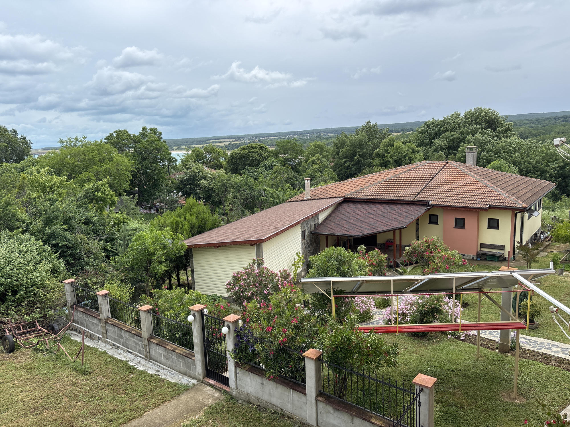 The image captures a panoramic view of a property in Rezovo, Bulgaria, under a cloudy sky. The scene is dominated by lush greenery, with a variety of trees and flowering bushes. In the foreground, a low, grey concrete wall with black metal fencing encloses a well-maintained lawn. To the left, a rustic, two-wheeled cart with a metal frame is parked on the grass.
A two-story house with a red-tiled roof stands prominently in the middle ground. The house has a yellow and pink facade, with dark-framed windows. A covered walkway or patio with a reddish-brown roof extends from the main structure. In the yard, a solar panel installation is visible, supported by metal poles. Several pink and purple flowering plants are in bloom around the house and yard.
Beyond the property, a dense forest stretches towards the horizon, interrupted by distant structures and fields. The expansive sky above is filled with dramatic, grey clouds, suggesting a possibility of rain. The overall impression is that of a tranquil, rural setting with a well-kept home surrounded by nature.