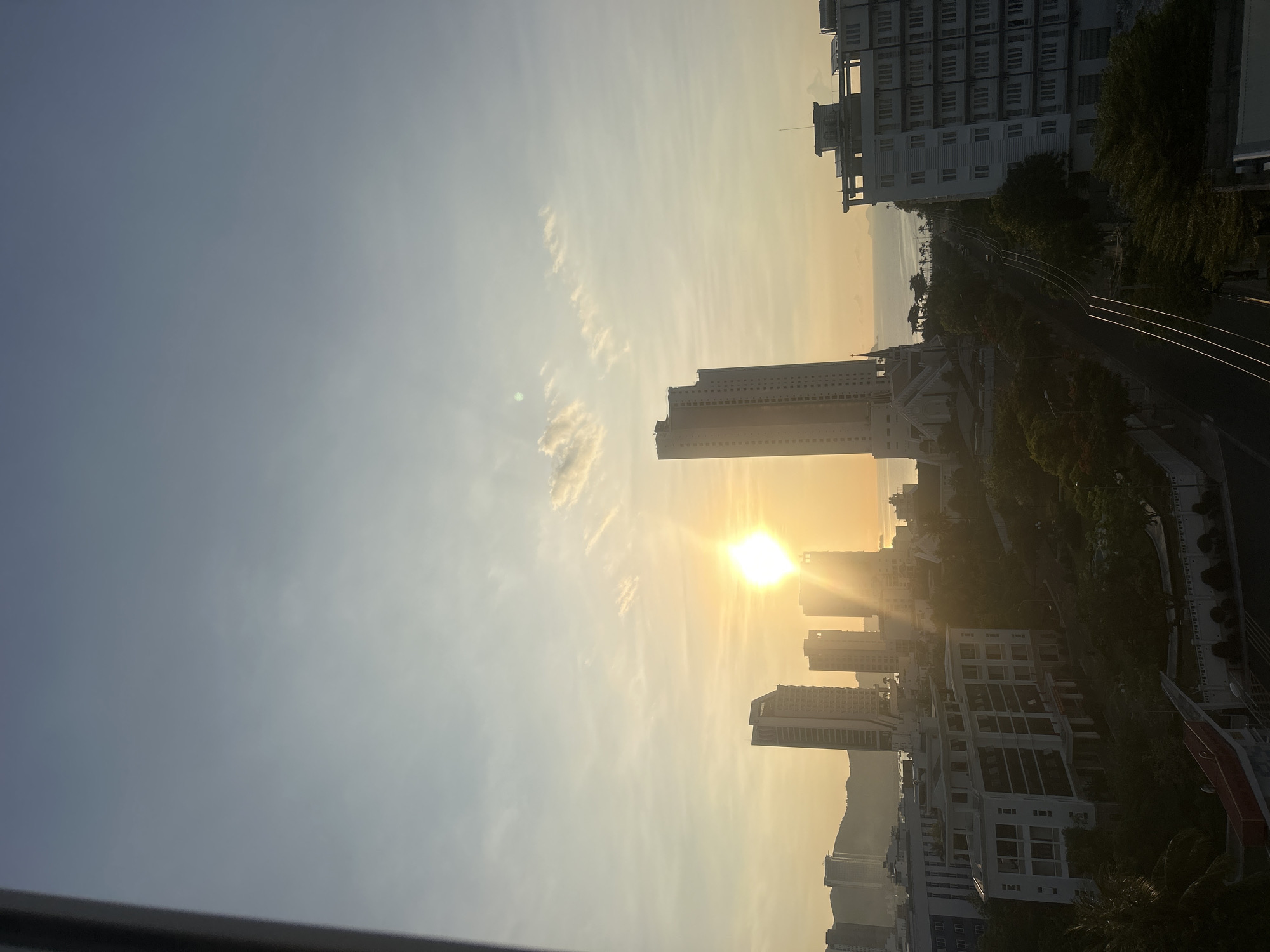 A horizontally oriented photograph captures a cityscape at sunrise or sunset, likely in the Bac Nha Trang Ward of Vietnam. The sun, a bright yellow orb, dominates the center of the frame, partially obscured by and casting a golden haze over several high-rise buildings. To the left of the sun, wispy clouds are visible against a blue sky that fades into a pale yellow near the horizon. The high-rise buildings are scattered, with varying heights and designs. To the right of the buildings are trees and a road.