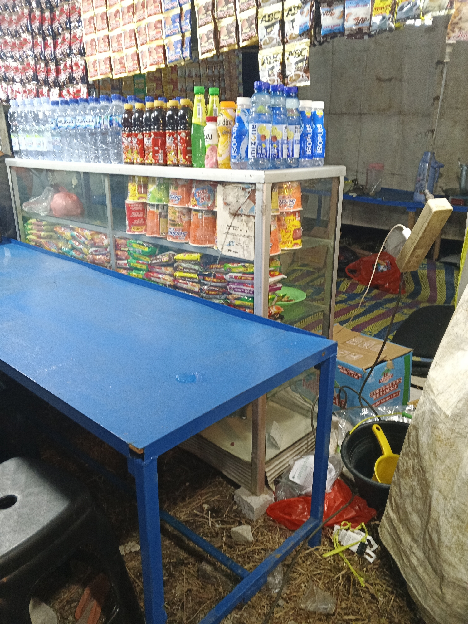 This image depicts an outdoor market stall in Sukabumi, Indonesia, likely at a roadside. The foreground features a bright blue, metal-framed table. Behind this table is a glass-fronted display cabinet filled with various packaged goods, including what appear to be instant noodles, snacks, and bottles of beverages. Above the cabinet, numerous small packages are hanging, displaying colorful labels that suggest food items or supplements.
To the right of the display cabinet, a black bucket with a yellow ladle is visible on the ground, alongside a red plastic bag and some debris. A large, rolled-up beige tarp or sack occupies the right side of the frame. Electrical wiring and an extension cord are also present, connecting to a power strip mounted on the wall or a vertical support. The ground is covered with dry grass or straw. The overall impression is of a small, modest retail operation, typical of informal markets in urban or semi-urban Indonesian settings.