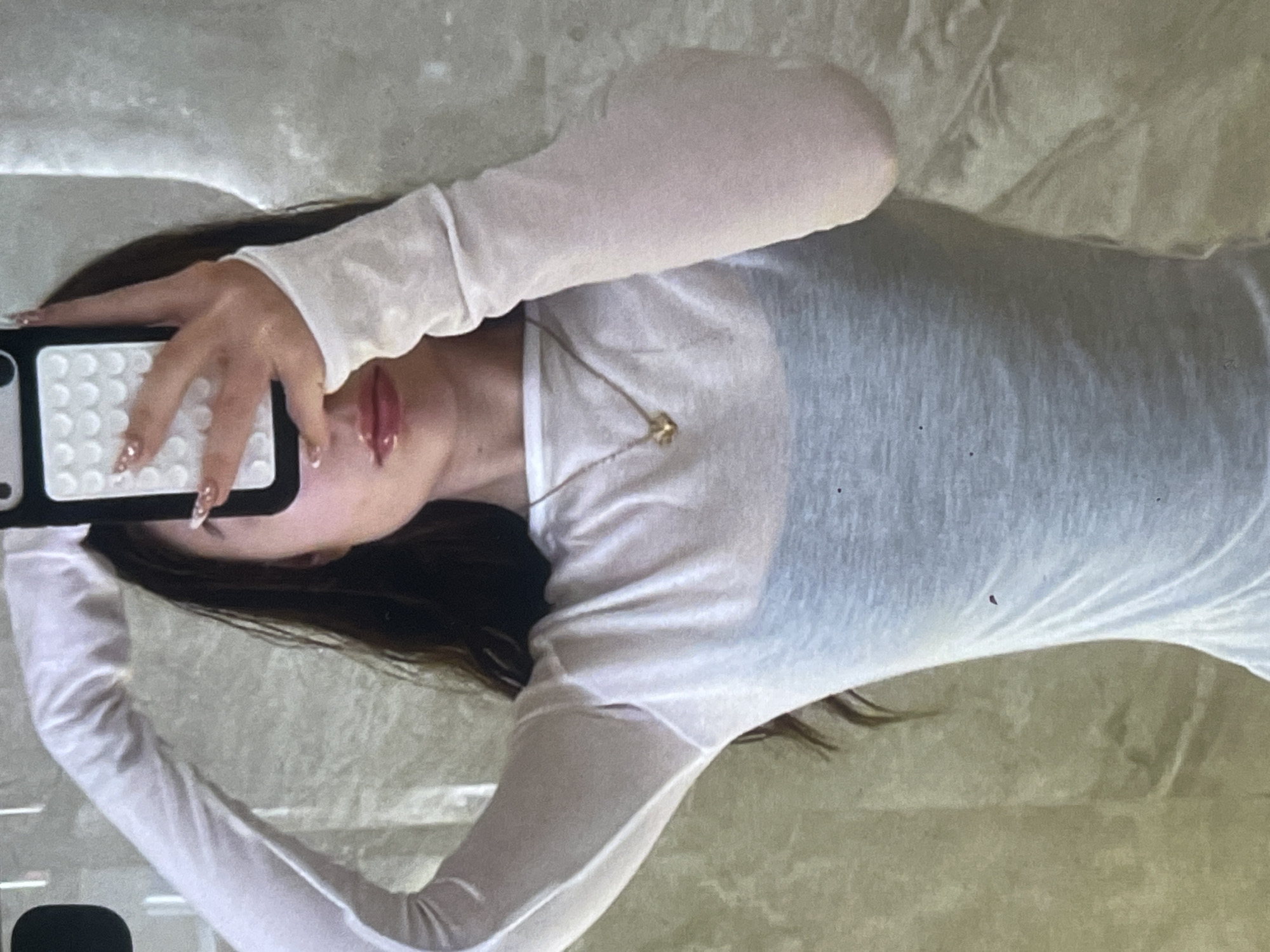 A person is captured in a mirror selfie, rotated 90 degrees clockwise, revealing their upper torso and head partially obscured by a smartphone. The individual, likely female, has long dark hair and wears a sheer, light-colored long-sleeved top over a darker undergarment, which is visible through the sheer fabric. A delicate gold-toned necklace with a small pendant rests on their chest.
The person holds a black smartphone with a distinctive white, grid-patterned case in their right hand. Their visible right hand shows long nails with intricate nail art, possibly involving glitter or an ombre effect. The lips are visible below the phone, appearing glossy and full, suggesting the application of lip gloss.
The background behind the person is a light, possibly textured wall or surface, reflecting warm, indoor lighting. There is no visible text in the image.
