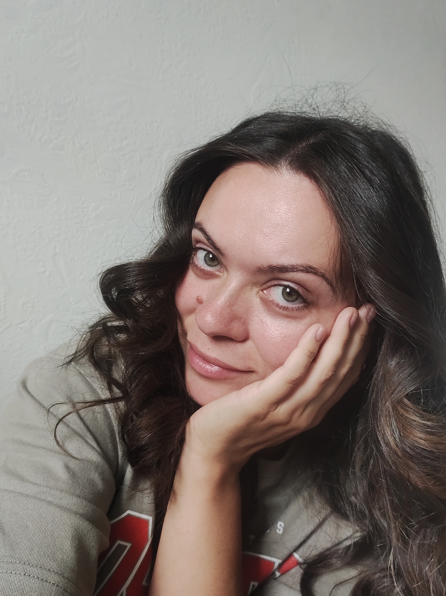 A woman is resting her head on her hand while looking towards the camera. She has long, dark, wavy hair and light-colored eyes. She is wearing a gray t-shirt with red and white lettering. The background is a textured white wall. The image appears to be taken indoors. No location in Chaikovsky, Russia is evident.