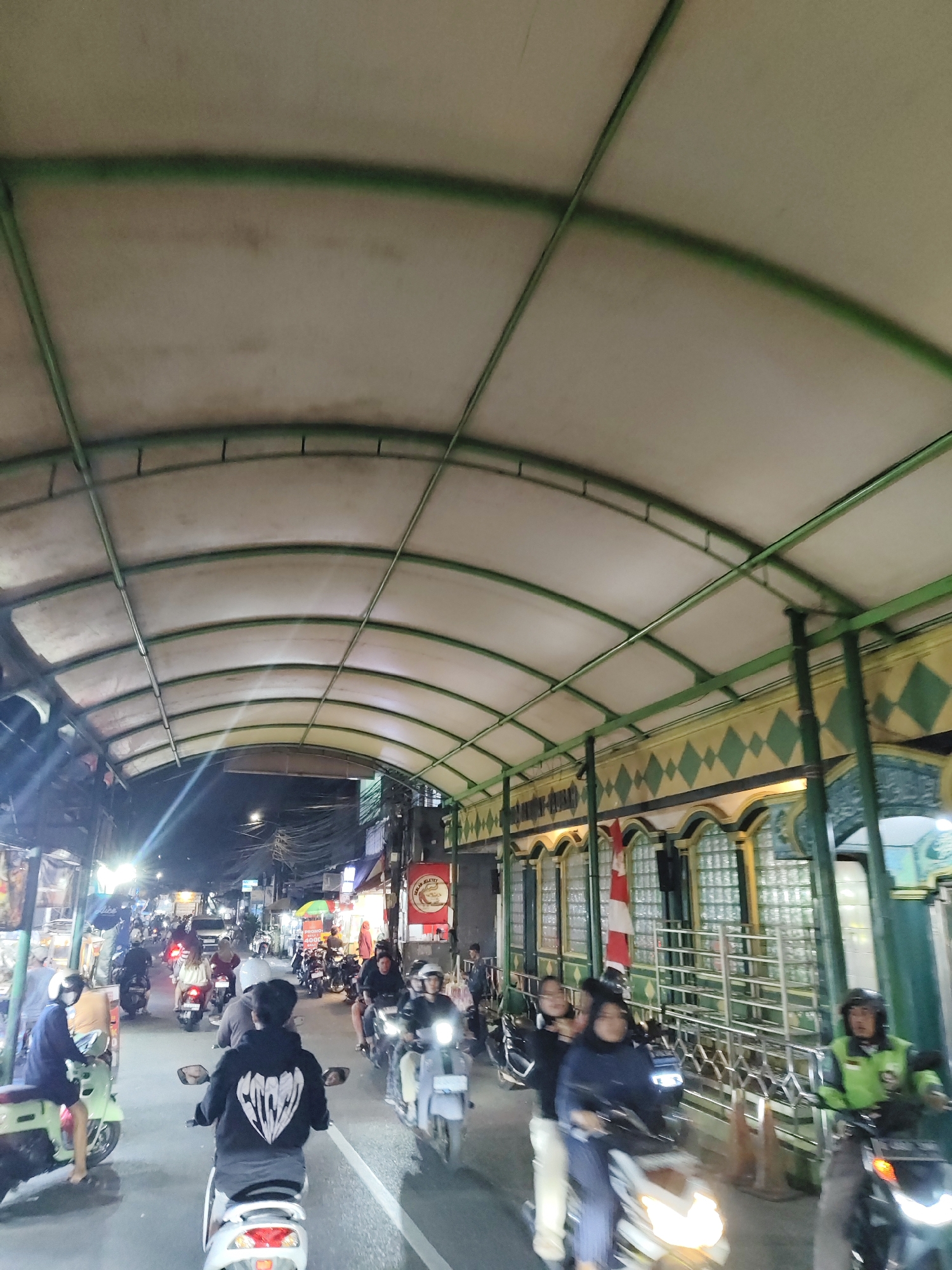This is a vertical, medium-shot, eye-level, photograph taken at night, showing a busy street scene in South Jakarta, Indonesia. The scene is dominated by a curved, translucent roof supported by green metal beams, casting a diffused light over the street below. Numerous motorcycles, ridden by people wearing helmets, are moving in both directions. The buildings lining the street are visible, featuring arched windows with glass blocks and decorative diamond patterns. Signs and lights suggest a commercial area, possibly a market or shopping street. The overall impression is one of vibrant nighttime activity in an urban environment.