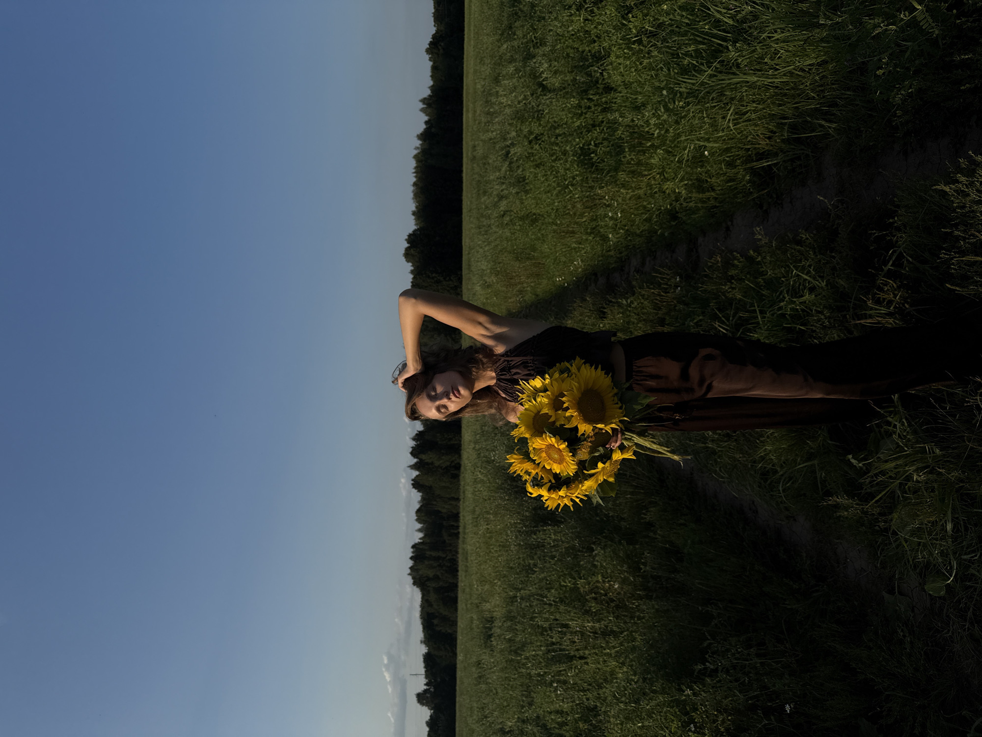 In Aksaitovo, Russia, a young woman stands in a tall grass field under a clear blue sky. She is holding a large bouquet of sunflowers. The woman is wearing a dark brown halter top and matching wide-leg pants. Her eyes are closed, and her right arm is raised, with her hand resting on her head. A dirt path runs through the field behind her, leading towards a dark line of trees in the distance. The lighting suggests it is late afternoon or early evening, with the sun casting shadows across the field.