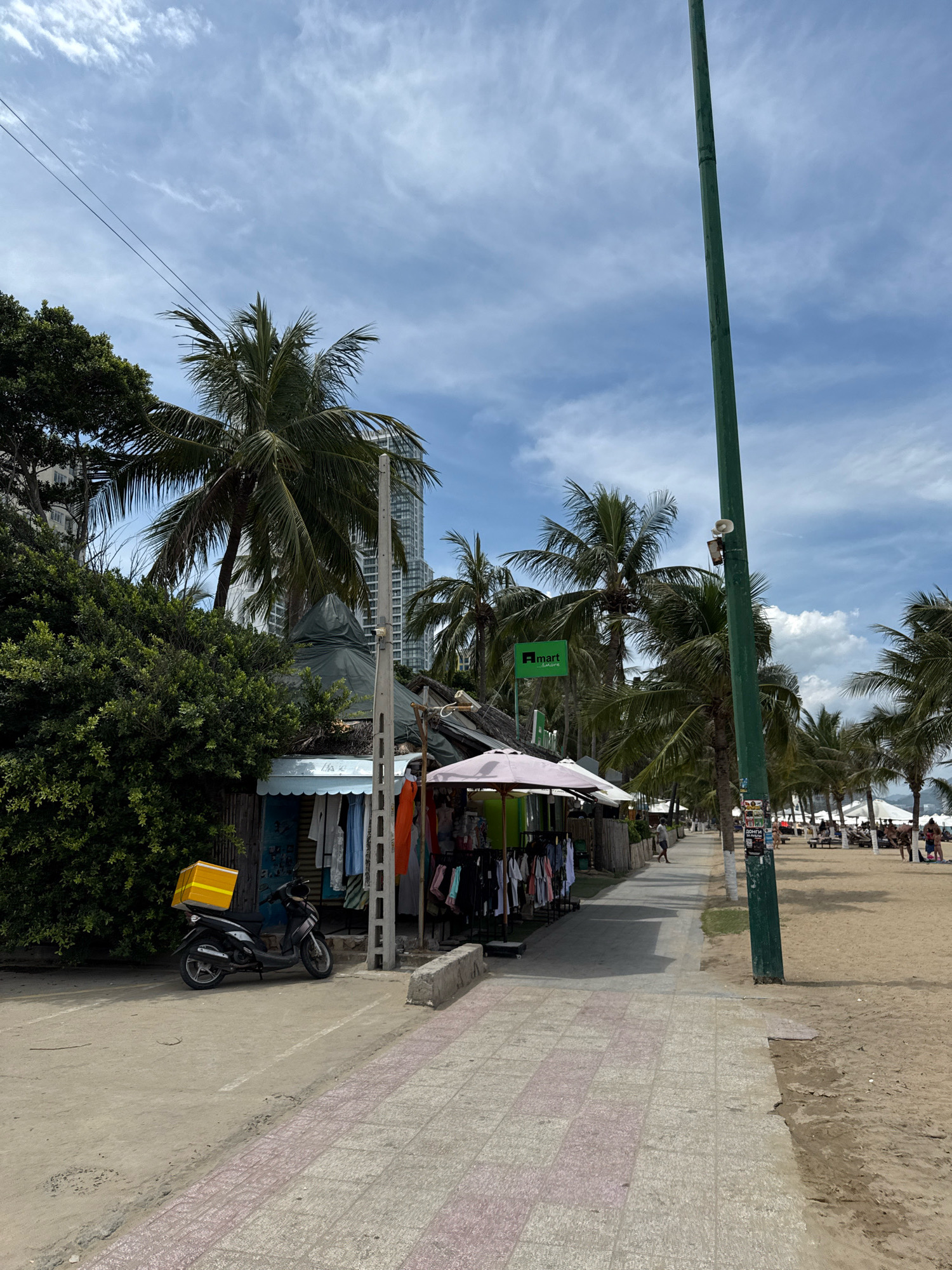 This image depicts a street scene in Nha Trang, Vietnam, likely near a beach. A paved walkway runs alongside a sandy beach, lined with palm trees and several small shops or stalls selling clothing. A black scooter with a yellow box on the back is parked to the left of the walkway, in front of a stall. In the background, people are seen relaxing on the beach, and tall buildings can be observed further inland. The sky is blue with scattered clouds, suggesting a sunny day. The overall atmosphere is one of a bustling tourist area.