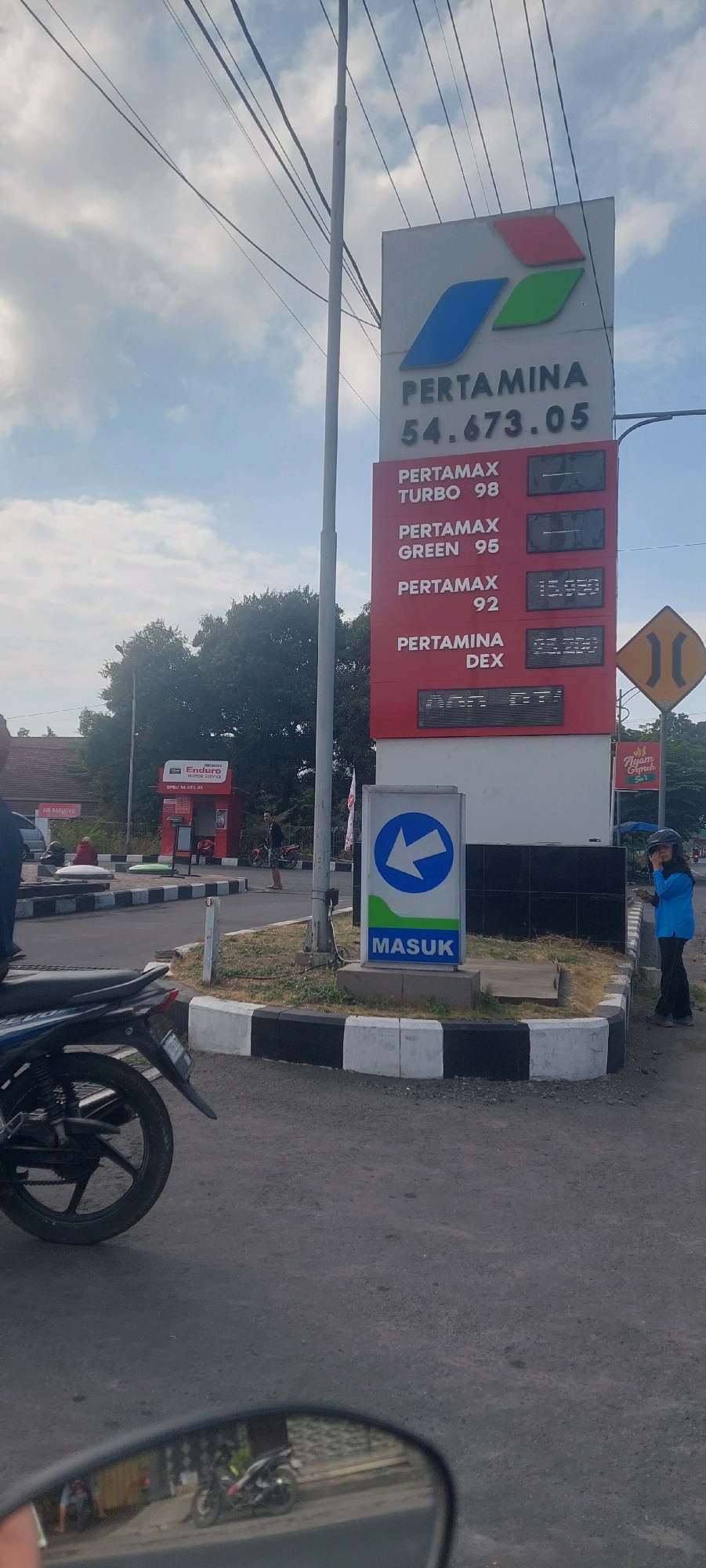 This is a street-level photograph taken in Lumajang, Indonesia, focusing on a Pertamina gas station price board. The price board displays prices for various fuel types: Pertamax Turbo 98, Pertamax Green 95, Pertamax 92, and Pertamina Dex. The prices shown are 15,950 for Pertamax 92 and 62,200 for Pertamina Dex. A sign with a blue arrow pointing left and the word "MASUK" (Indonesian for "enter") is visible in the foreground. In the background, there is a red structure with a "Enduro Motor Service" sign. A motorcycle is parked in the foreground, and its reflection is visible in a mirror in the lower portion of the image, showing a street scene with buildings and another motorcycle. The sky is overcast with scattered clouds. Two individuals are visible in the mid-ground, one standing near a red structure and another standing by the side of the road wearing a blue shirt and black pants, holding a phone. A yellow directional sign with two parallel lines is also present. The environment appears to be an urban or semi-urban setting with a paved road and sidewalks.
