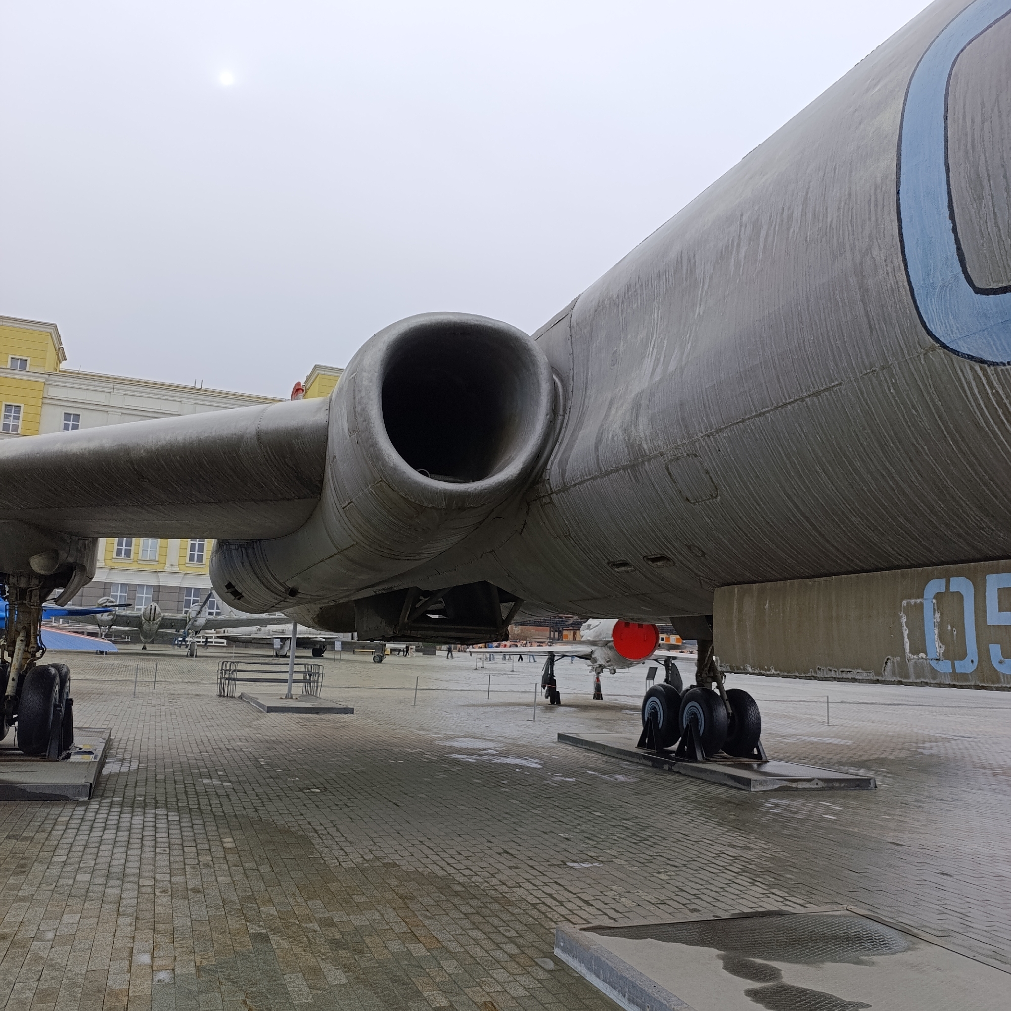 The image presents an outdoor scene featuring a large, weathered grey aircraft on display, likely at an aviation museum in Verkhnyaya Pyshma, Russia. The perspective is from a low angle, looking up at the underside and right side of the aircraft.
The primary subject is the immense fuselage and wing of a large aircraft, possibly a bomber or transport plane, seen from its starboard side. A prominent engine nacelle, which appears to house a jet engine, extends from the wing. The aircraft's landing gear, featuring multiple dark tires, rests on a concrete platform. The metallic surface of the aircraft shows significant weathering, with numerous dark streaks running vertically down the fuselage, indicating exposure to the elements. A large, stylized blue "C" shape and the partial number "05" are visible painted on the side of the fuselage near the tail section.
In the background, several other aircraft of various sizes and types are visible on display, further confirming the setting as an aviation exhibition. To the left, a multi-story building with a distinctive yellow facade and numerous white-framed windows stands prominently. Throughout the background, several people are scattered, walking on the paved ground, suggesting visitors to the museum.
The ground is covered with grey, rectangular paving stones, exhibiting patches of wetness that contrast with drier areas, possibly from recent rain or melting snow. The sky is uniformly overcast, casting a soft, diffuse light over the scene. A bright, indistinct circle in the upper sky indicates the sun obscured by clouds. No specific activity beyond observation and walking is evident among the people. The overall impression is one of quiet contemplation at an outdoor museum on a cool, cloudy day. There are no discernible emotions displayed.
