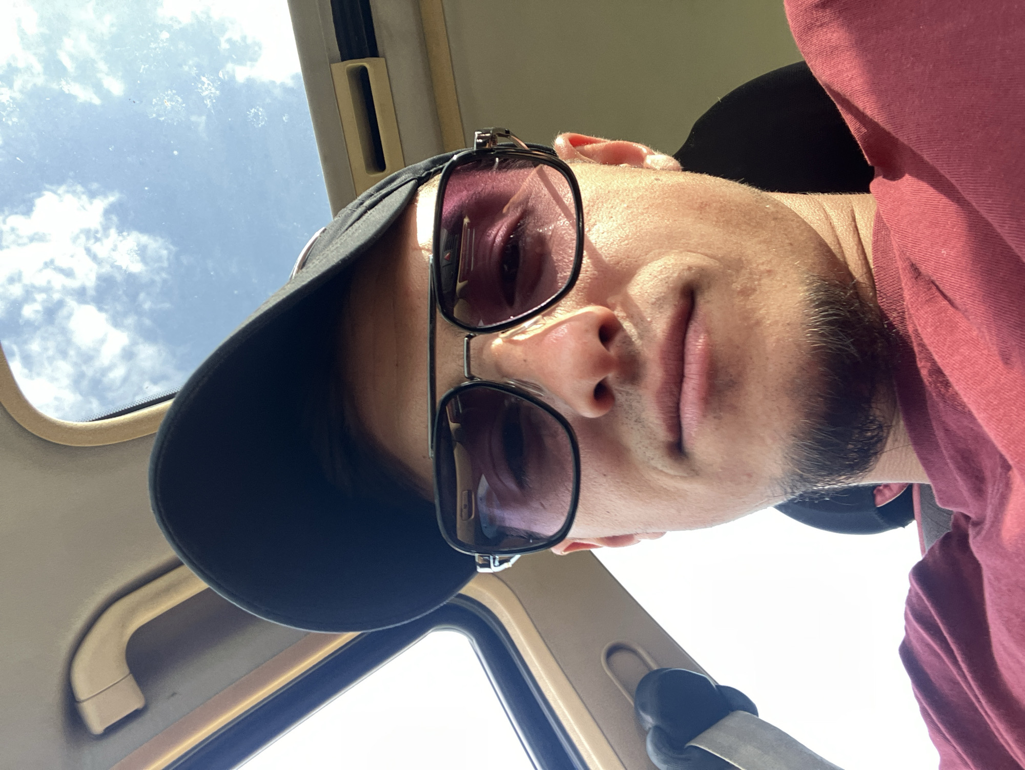 A person with a black cap and sunglasses is inside a car, looking down towards the camera. The car's interior is visible, with the sky and clouds seen through the sunroof. The person is wearing a maroon shirt. The location cannot be confirmed as Krastevich, Bulgaria from the image.