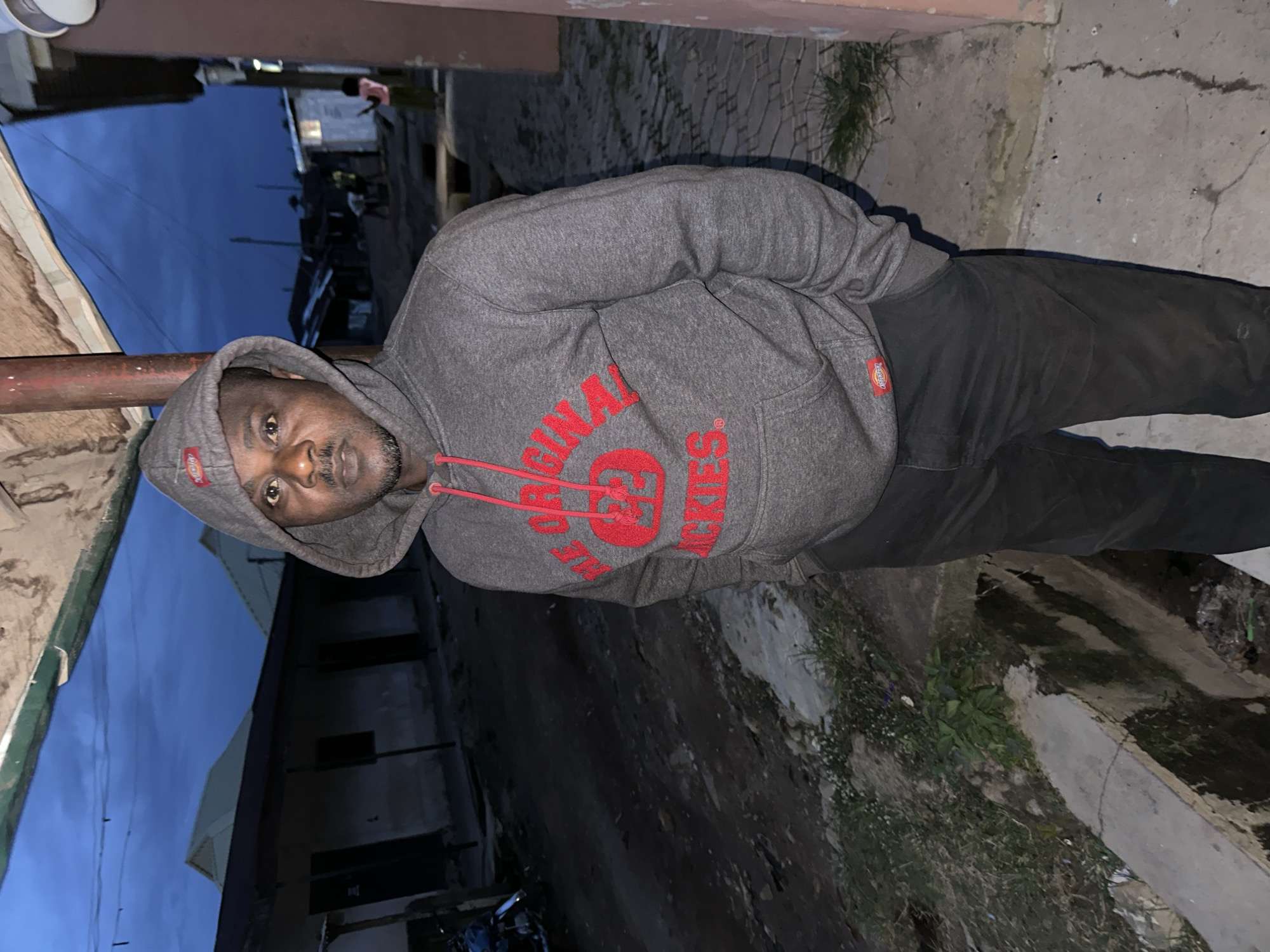 The image depicts a man wearing a gray hoodie with red lettering and black pants, standing in what appears to be an outdoor setting at dusk. The man is facing the camera, with his hands in his pockets. The background shows a street scene with buildings, some of which are illuminated. Electrical wires are visible overhead, and there is a motorcycle parked on the left side of the frame. The ground is paved with what looks like cobblestones in some areas and dirt in others. A concrete drainage channel is visible in the foreground. The sky is dark blue, suggesting it is either early evening or late evening. The overall impression is a candid street photograph taken in a residential or semi-commercial area, possibly in Narayi, Nigeria, given the architectural style and the presence of what appear to be local structures.
