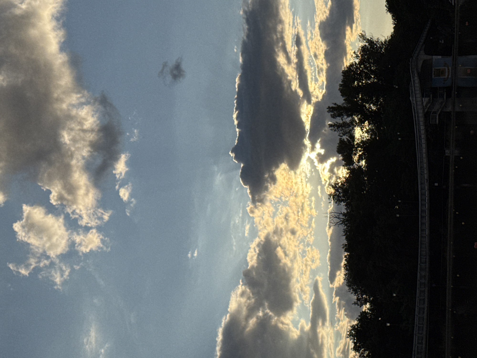The image captures a dramatic sky with scattered clouds at sunset, casting golden light and shadows over a silhouette of trees and a bridge. The sky transitions from a pale blue at the top to brighter, warmer tones near the horizon where the sun is setting. The clouds are a mix of dark, heavy masses and lighter, illuminated wisps.
Below the sky, a dense line of dark trees forms a silhouette against the vibrant sky. A pedestrian bridge with a railing and some architectural features, possibly a small building or structure beneath it, is visible in the mid-ground. The bottom of the image shows a dark, reflective surface, likely water, with some indistinct shapes on it. Small lampposts are visible within the treeline, suggesting a park or waterfront area.
This scene appears to be the area around the Rowing Canal in Plovdiv, Bulgaria. The specific viewpoint is likely from the water's edge, looking towards the treeline and the bridge that spans part of the canal. The time of day is dusk, with the setting sun creating a picturesque, moody atmosphere. There are no people clearly visible in the frame.