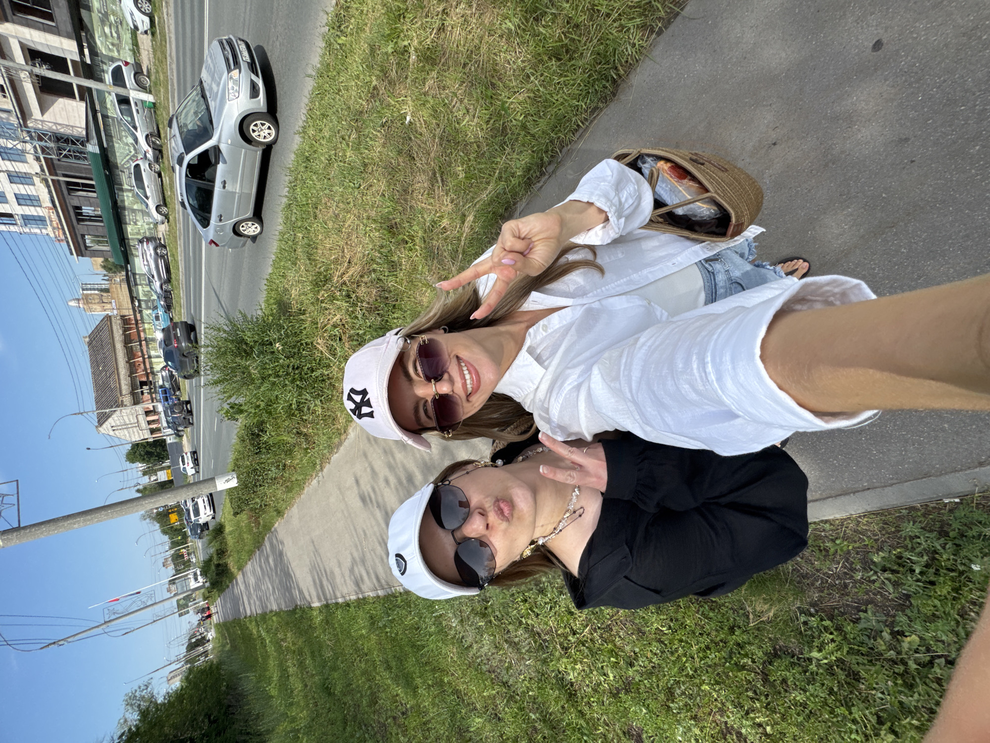 Two women are taking a selfie on a sidewalk next to a street. The woman on the left is wearing a black long-sleeved shirt, a white baseball cap, and sunglasses. She is making a duck face and holding up two fingers in a peace sign. The woman on the right is wearing a white button-down shirt, denim shorts, and a pink baseball cap. She is smiling and also holding up two fingers in a peace sign. She has a woven tote bag at her side. The background shows a street with cars, buildings, and trees under a clear blue sky.